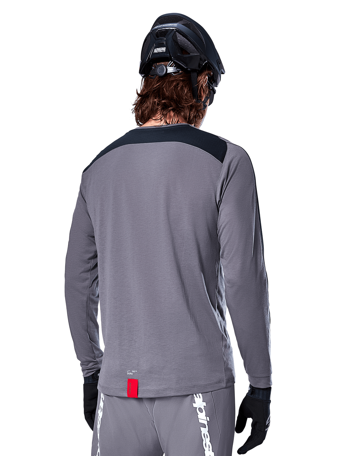 Alpinestars A-Dura Dri Astar Langarmtrikot, dunkelgraues MTB-Trikot von hinten gezeigt, mit schwarzem Schulterbereich und kleinem roten Markenlabel, kombiniert mit schwarzem Mountainbike-Helm und Handschuhen.