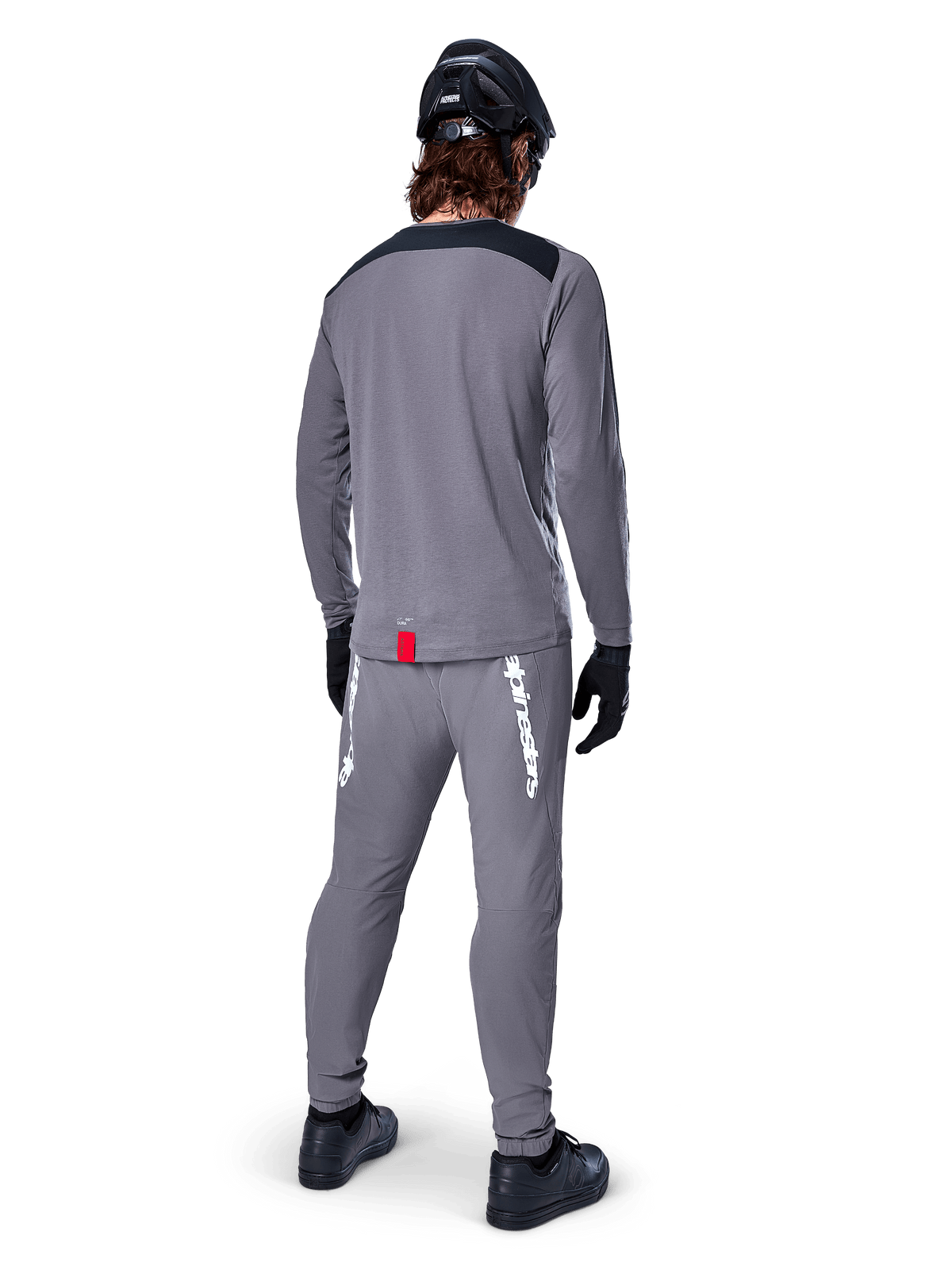 Alpinestars A-Dura Dri Astar Langarmtrikot, Dunkelgrau, Rückansicht eines Fahrers mit passender Hose und schwarzem Helm, mit schwarzen Schultereinsätzen und einem kleinen roten Etikett am Saum, technische Offroad-Radbekleidung
