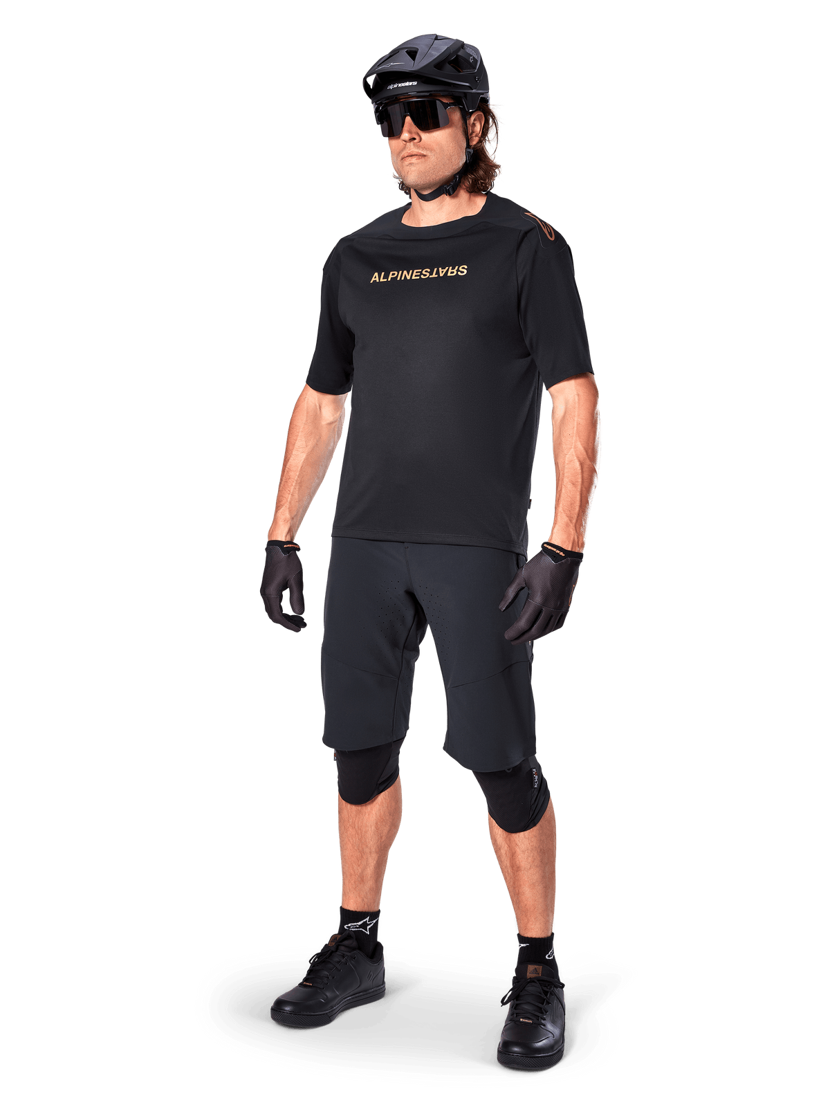 Maillot à manches courtes Alpinestars A-Aria Polartec Switch, noir, avec marquage doré effet miroir sur la poitrine, porté par un cycliste avec un short de cyclisme noir, un casque et des genouillères, équipement technique de VTT pour la performance tout-terrain