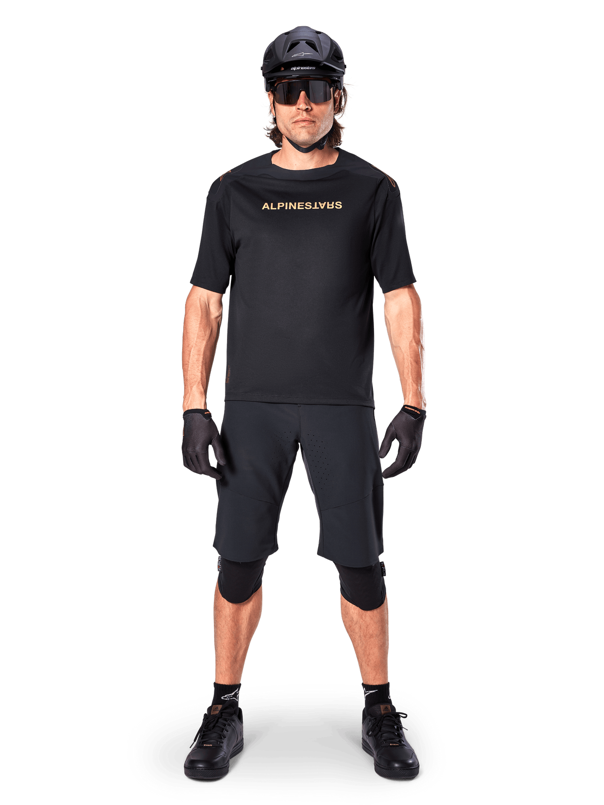 Alpinestars A-Aria Polartec Switch Jersey, maillot de VTT à manches courtes, noir avec lettrage beige, porté avec un équipement de VTT noir coordonné comprenant casque, short et genouillères, vêtement de cyclisme tout-terrain haute performance