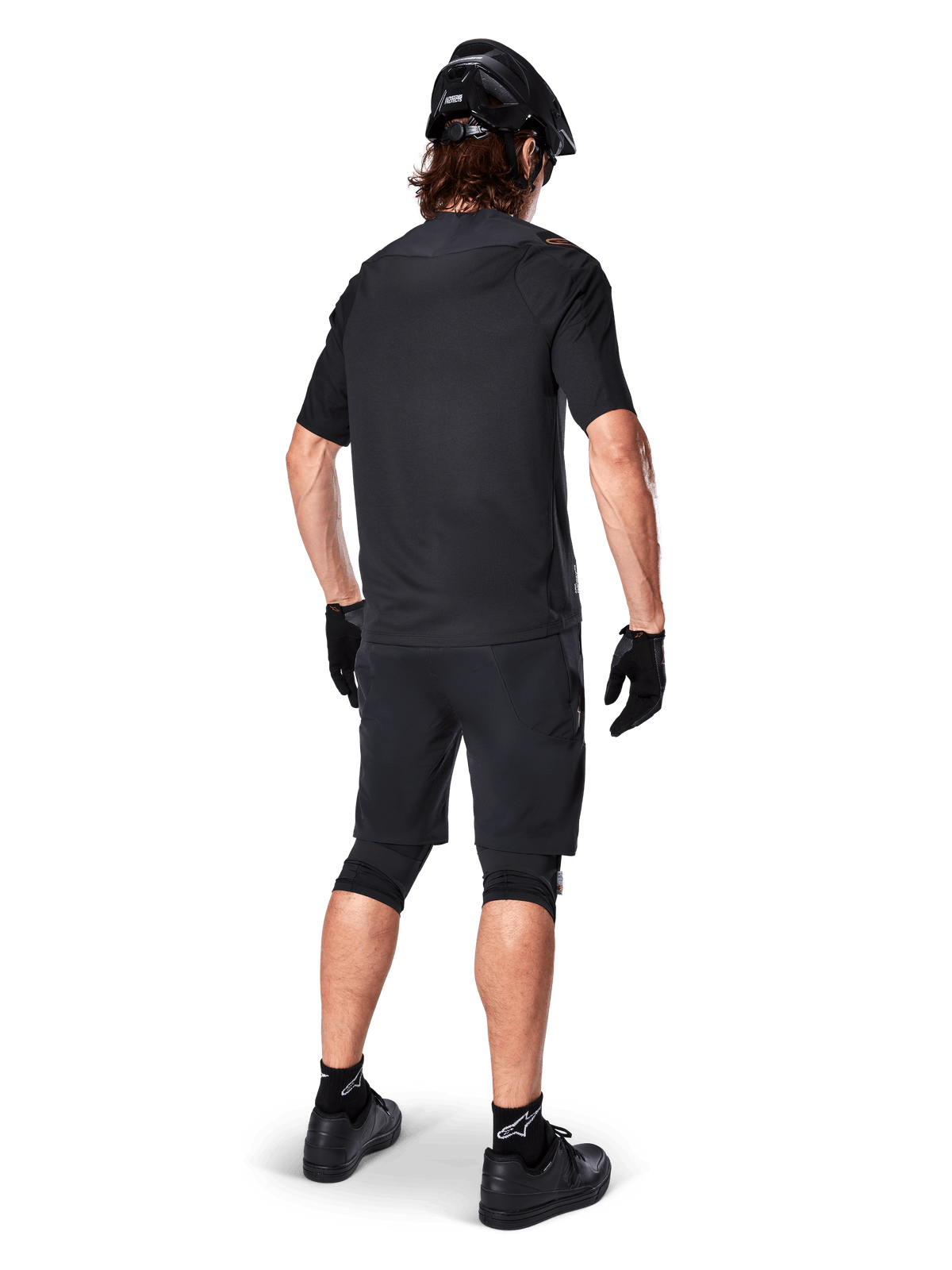 Cycliste de VTT de dos portant le maillot Alpinestars A-Aria Polartec Switch noir, maillot de VTT à manches courtes, associé à un short de VTT noir, un casque et des gants noirs, tenue de performance coordonnée pour la pratique sur sentiers tout-terrain
