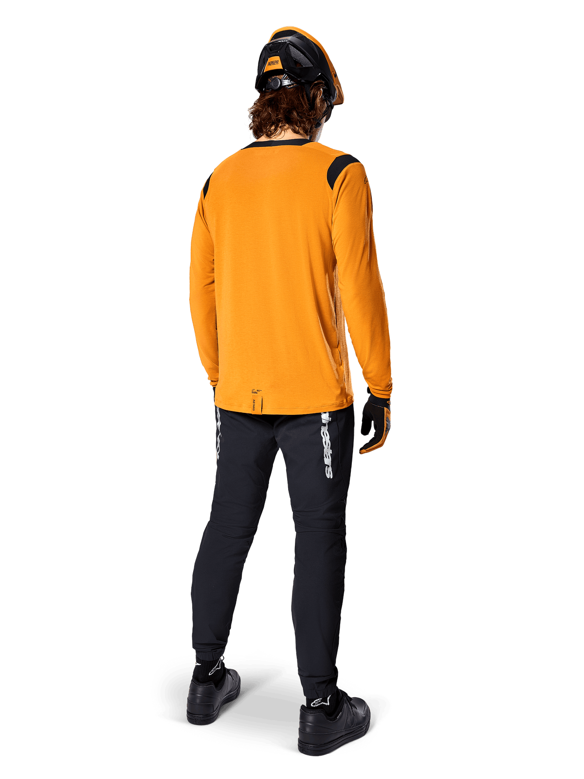 Vététiste vu de dos portant le maillot à manches longues Alpinestars A-Dura Dri Wool en Dark Gold, un pantalon technique noir avec des logos blancs et un casque noir et orange assorti, équipement de VTT haute performance.