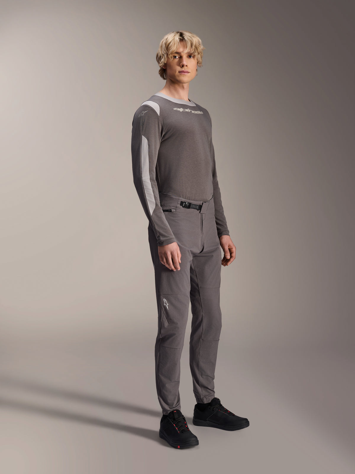 Alpinestars A-Dura Elite Pants, mountainbikebroek, donkergrijs, gedragen met een bijpassende jersey met lange mouwen en zwarte schoenen, met een ratelsluiting aan de voorzijde en duurzame Cordura-stof voor trail- en enduro-ritten.