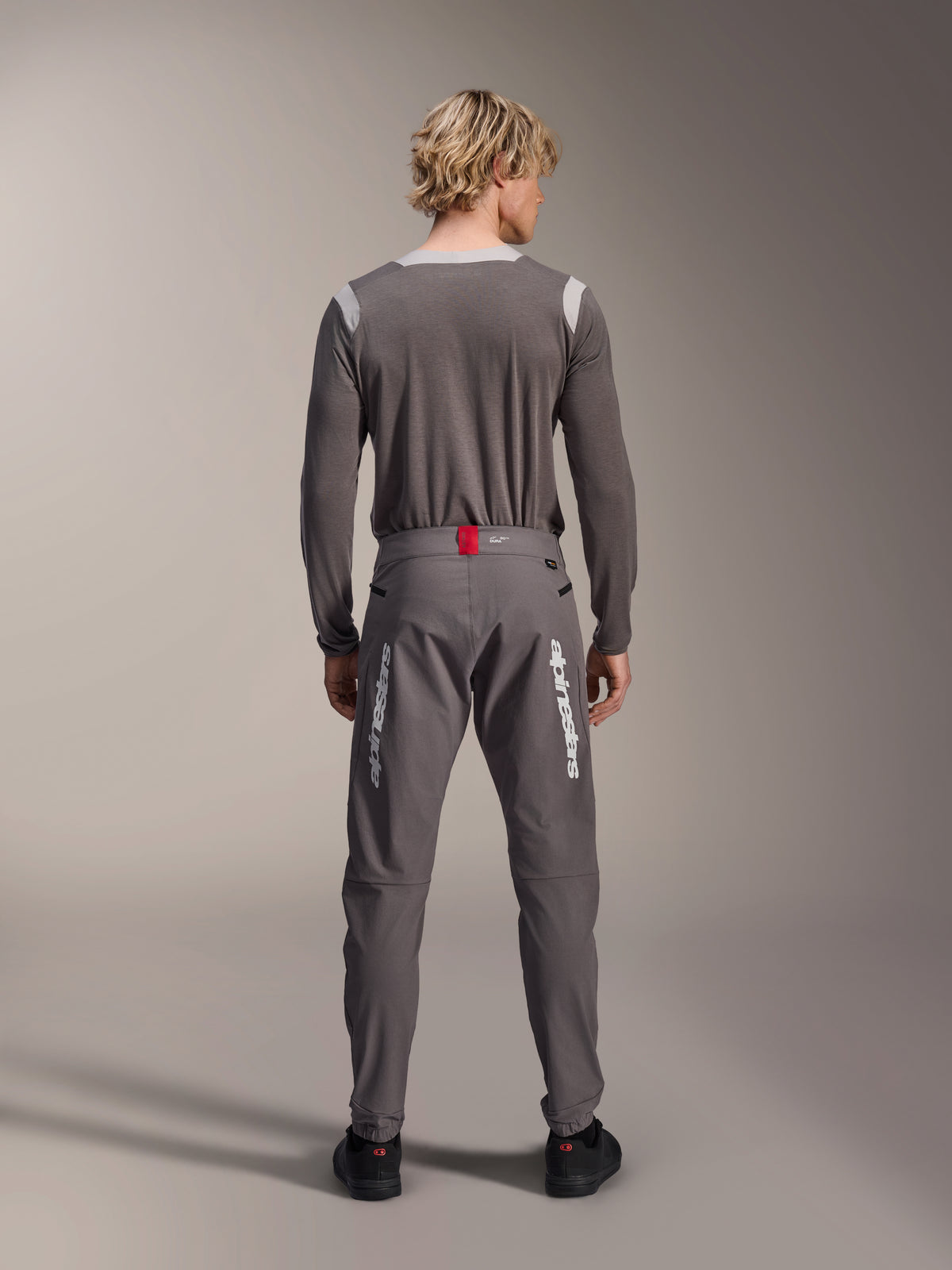 Alpinestars A-Dura Elite Pants in donkergrijs, model van achteren gezien met bijpassende grijze jersey met lange mouwen, witte Alpinestars-logo's op de pijpen, rood accent op de tailleband, zwarte schoenen, professionele MTB-uitrusting