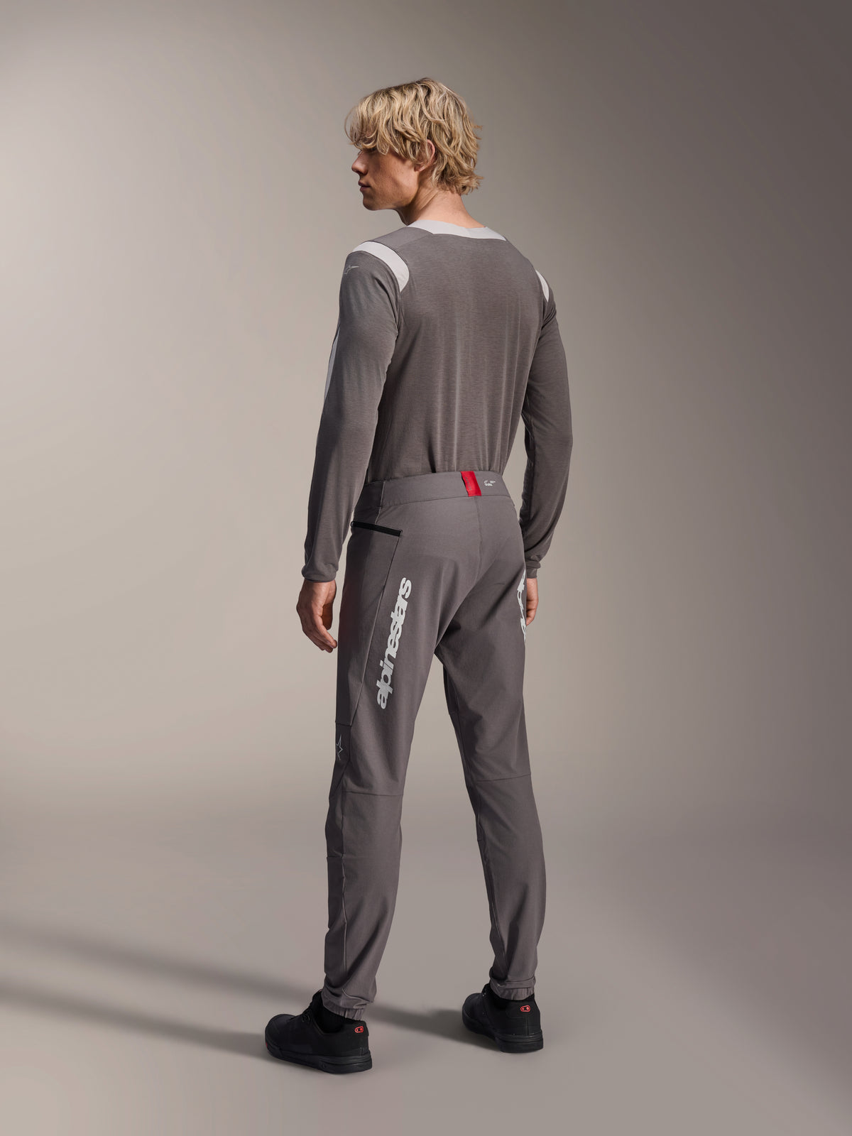 Model van achteren gezien in Alpinestars MTB-kleding, grijze A-Dura Dri Wool Jersey met lange mouwen, donkergrijze A-Dura Elite Pants met witte verticale branding en rood detail op de tailleband, gecoördineerde fietsoutfit voor mountainbiken