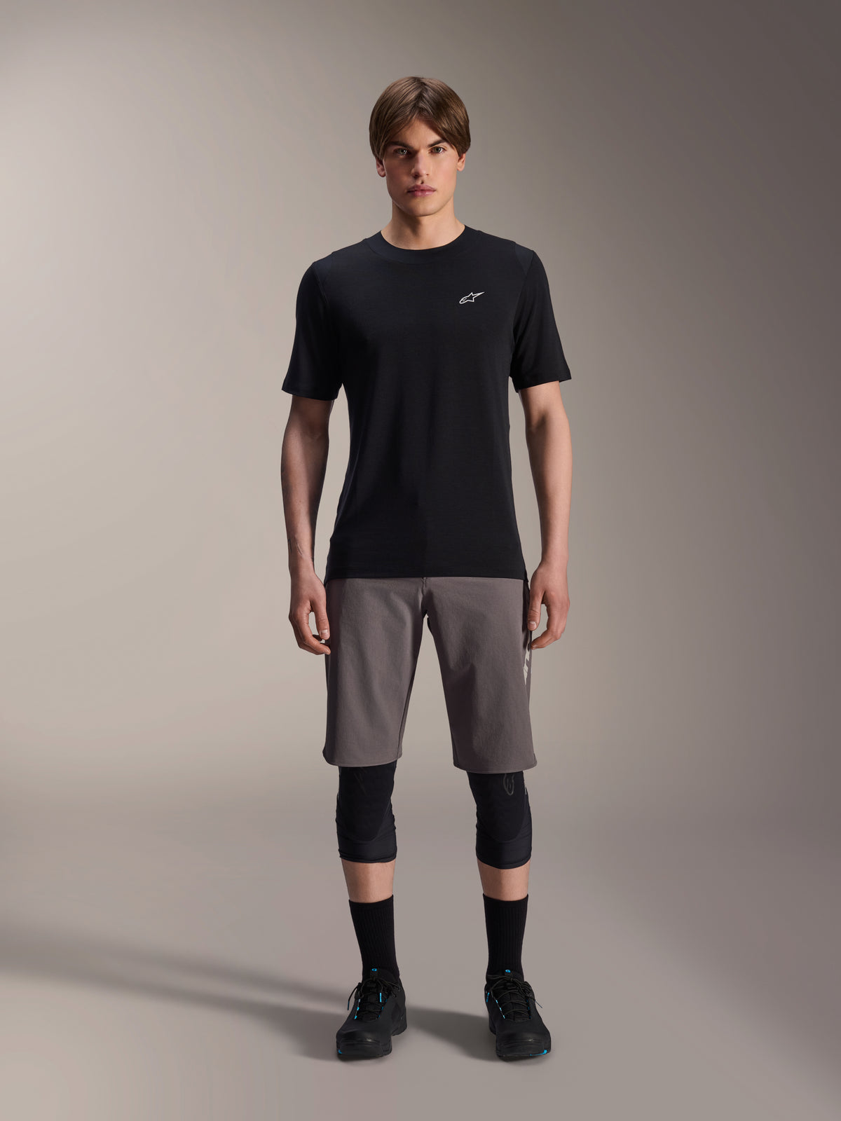 Alpinestars A-Dura Dri Wool Kurzarmtrikot, Schwarz, getragen von einem Model mit grauen Mountainbike-Shorts, schwarzen Knieprotektoren und schwarzen Fahrradschuhen, minimalistisches Design mit einem kleinen weißen Astars-Logo auf der Brust, funktionelle Performance-MTB-Bekleidung für Trail-Fahrten.