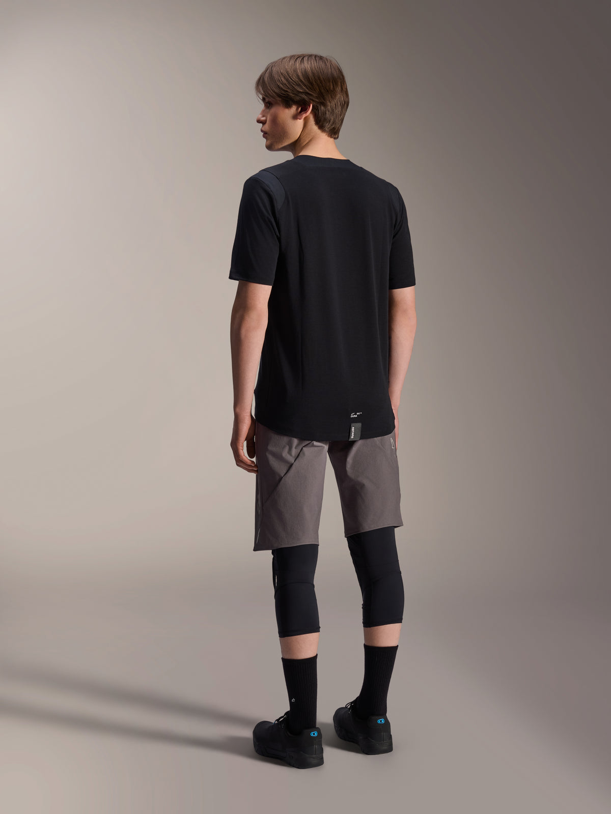 Alpinestars A-Dura Dri Wool Kurzarmtrikot, Schwarz, Rückansicht am Modell, getragen mit grauen Mountainbike-Shorts und schwarzen Knieprotektoren, mit verlängertem Rückensaum und dezentem Logo, technische MTB-Bekleidung