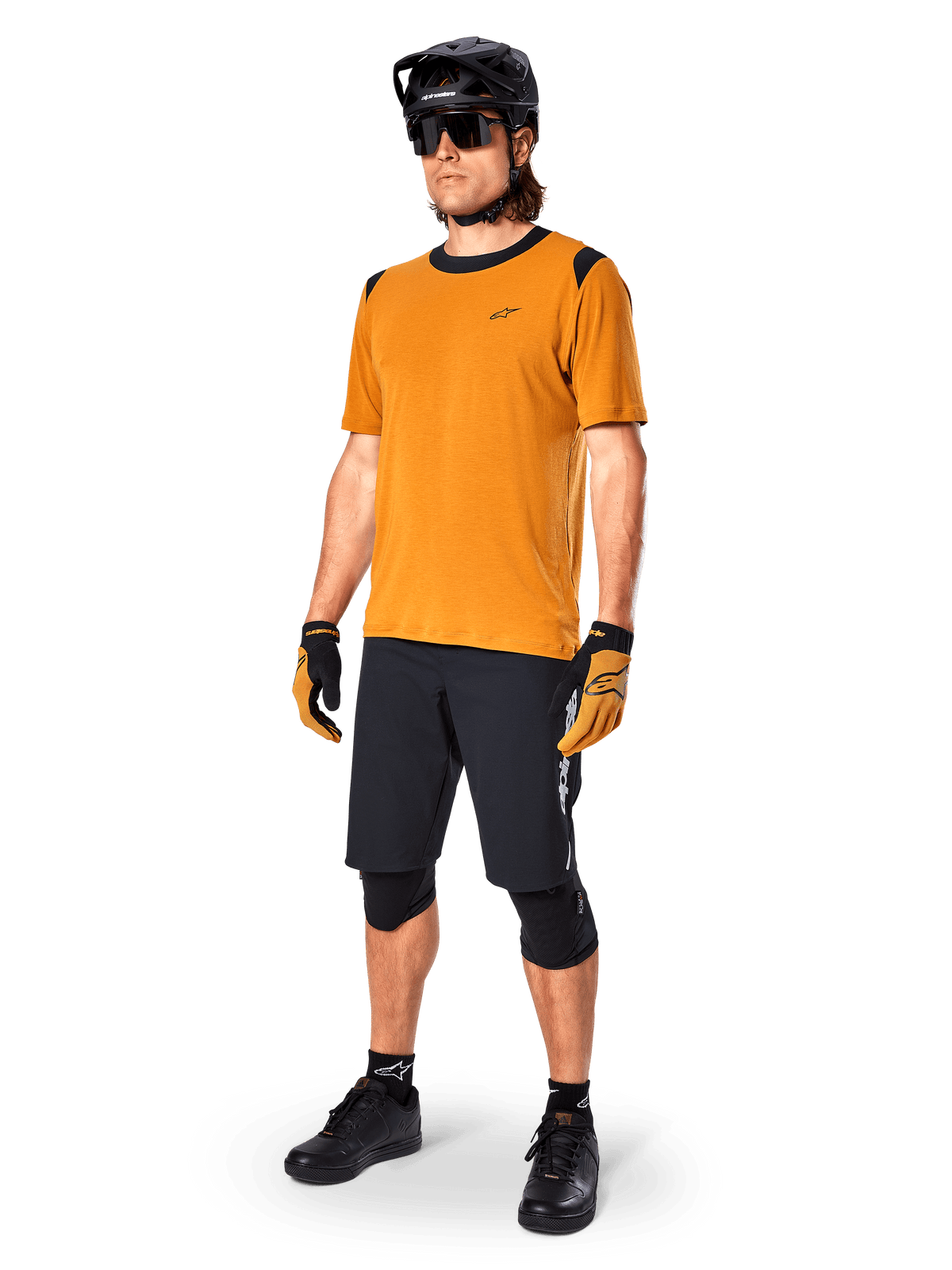 Modèle masculin portant le maillot Alpinestars A-Dura Dri Wool à manches courtes en Dark Gold, maillot de VTT avec accents noirs aux épaules et au col, associé à un short de VTT noir, un casque noir et des lunettes de soleil, équipement technique de cyclisme tout-terrain