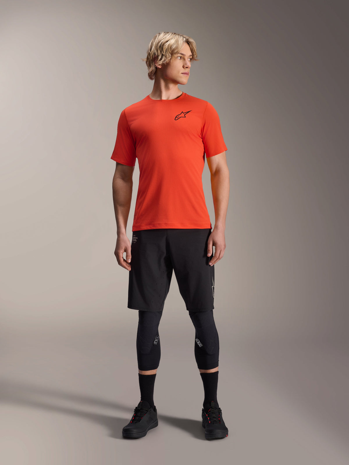 Alpinestars A-Aria Astar Kurzarmtrikot, Spicy Orange, getragen von einem Model mit schwarzen Mountainbike-Shorts und Knieschützern, leichte und atmungsaktive Performance-Radsportbekleidung für MTB