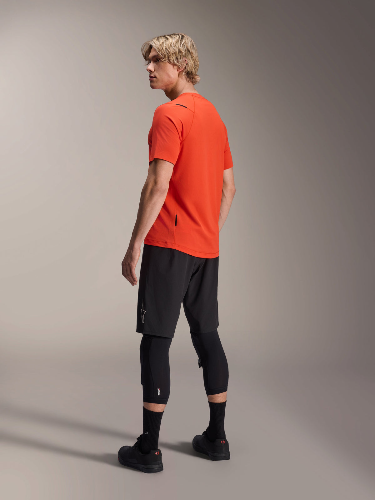 Alpinestars A-Aria Astar Kurzarmtrikot, Spicy Orange, Dreiviertel-Rückansicht am Modell mit Blick über die Schulter, kombiniert mit schwarzen MTB-Shorts und Knieprotektoren, Performance-Mountainbike-Bekleidung
