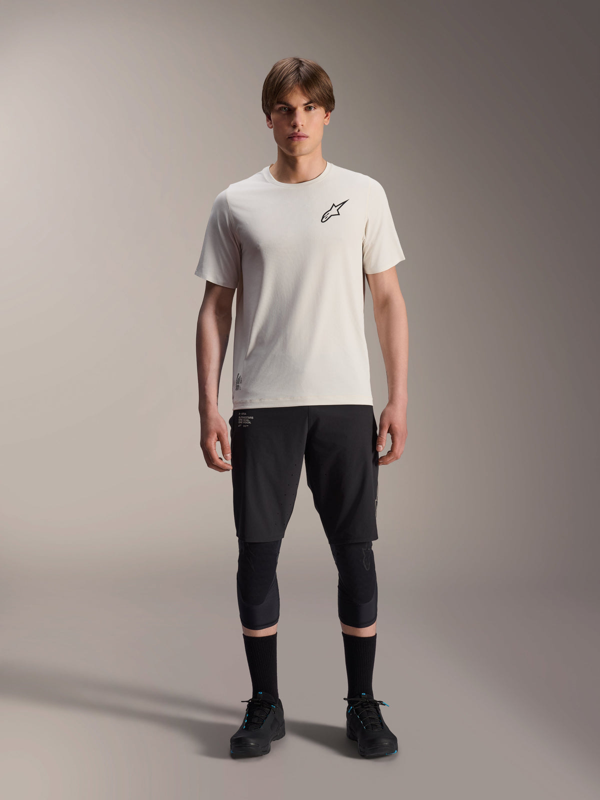 Modèle masculin portant le maillot à manches courtes Alpinestars A-Aria Astar en Écru, associé à un short de VTT noir, des genouillères et des chaussures de cyclisme noires, avec un logo étoile noir minimaliste sur la poitrine, conçu pour la pratique du trail tout-terrain.
