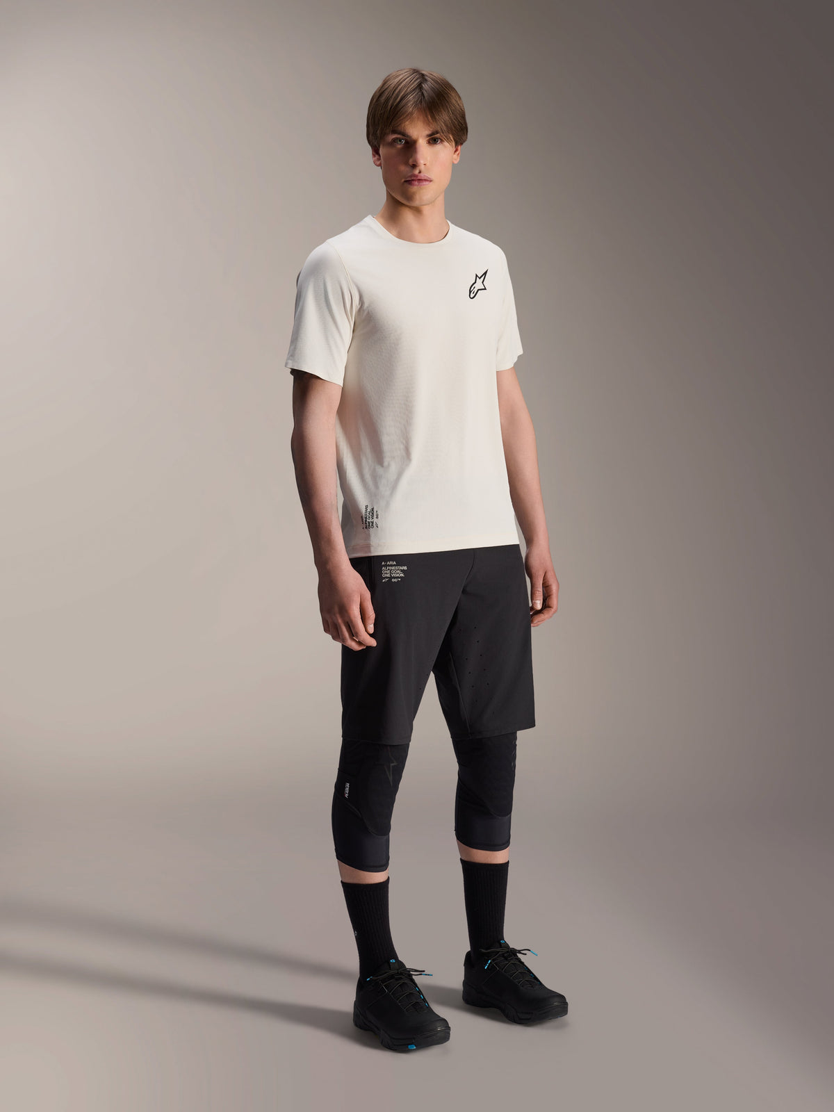 Maillot à manches courtes Alpinestars A-Aria Astar, écru, avec logo noir sur la poitrine, porté avec un short de cyclisme noir et des genouillères, équipement VTT léger et performant