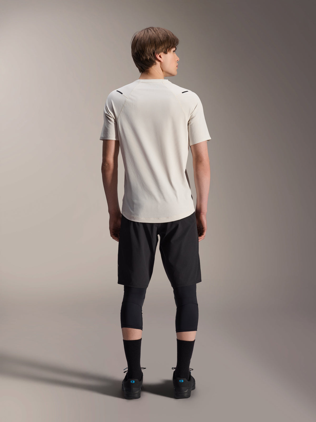 Modèle masculin vu de dos portant le maillot à manches courtes Alpinestars A-Aria Astar en Écru, associé à un short de VTT noir et des leggings de protection, avec un pan arrière allongé et une coupe performance pour la conduite sur sentiers tout-terrain.