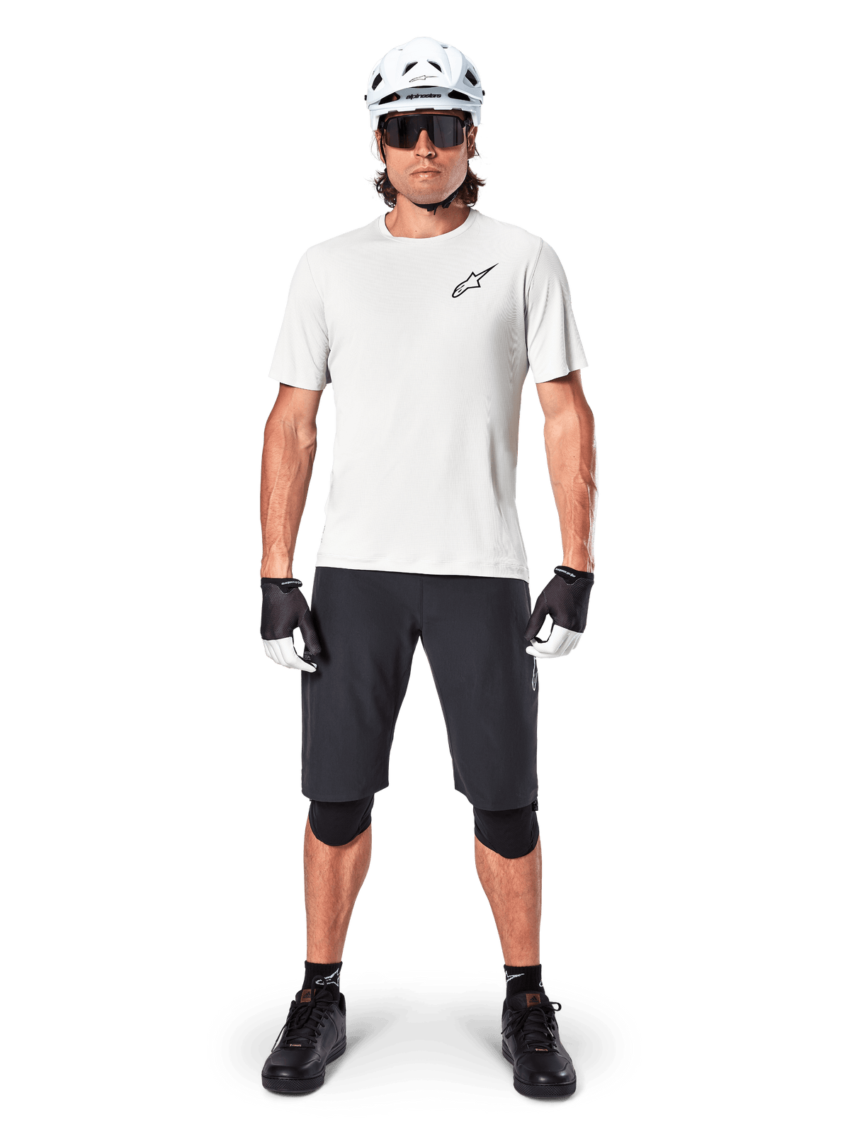 Mannelijke fietser die het Alpinestars A-Aria Astar Jersey met korte mouwen in White Dove draagt, voorzien van een zwart Astars-logo op de borst, gecombineerd met een zwarte mountainbike short, witte helm, zonnebril, zwarte kniebeschermers en handschoenen, complete off-road fietsoutfit.