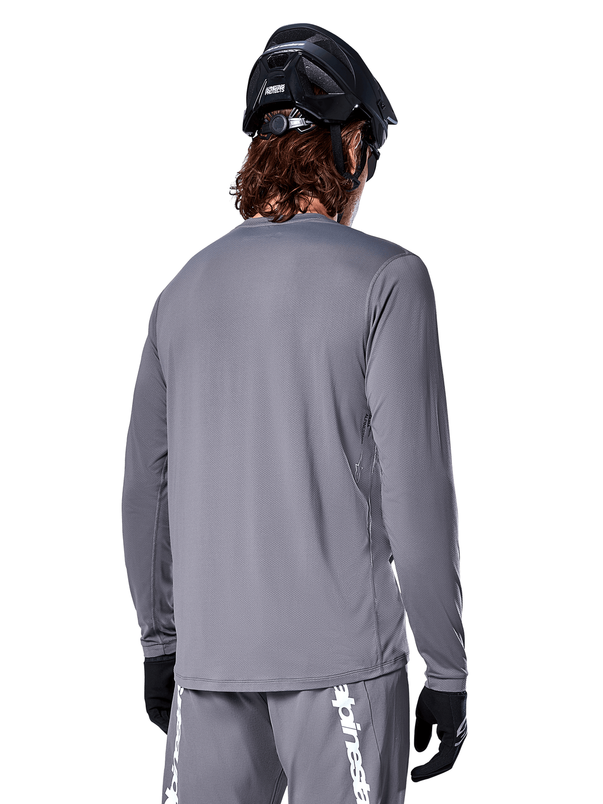 Maillot à manches longues Alpinestars A-Dura Rocker en gris foncé, vue arrière d'un cycliste de VTT, avec un short gris assorti arborant un logo blanc, un casque de VTT noir et des gants noirs, équipement de cyclisme tout-terrain professionnel.