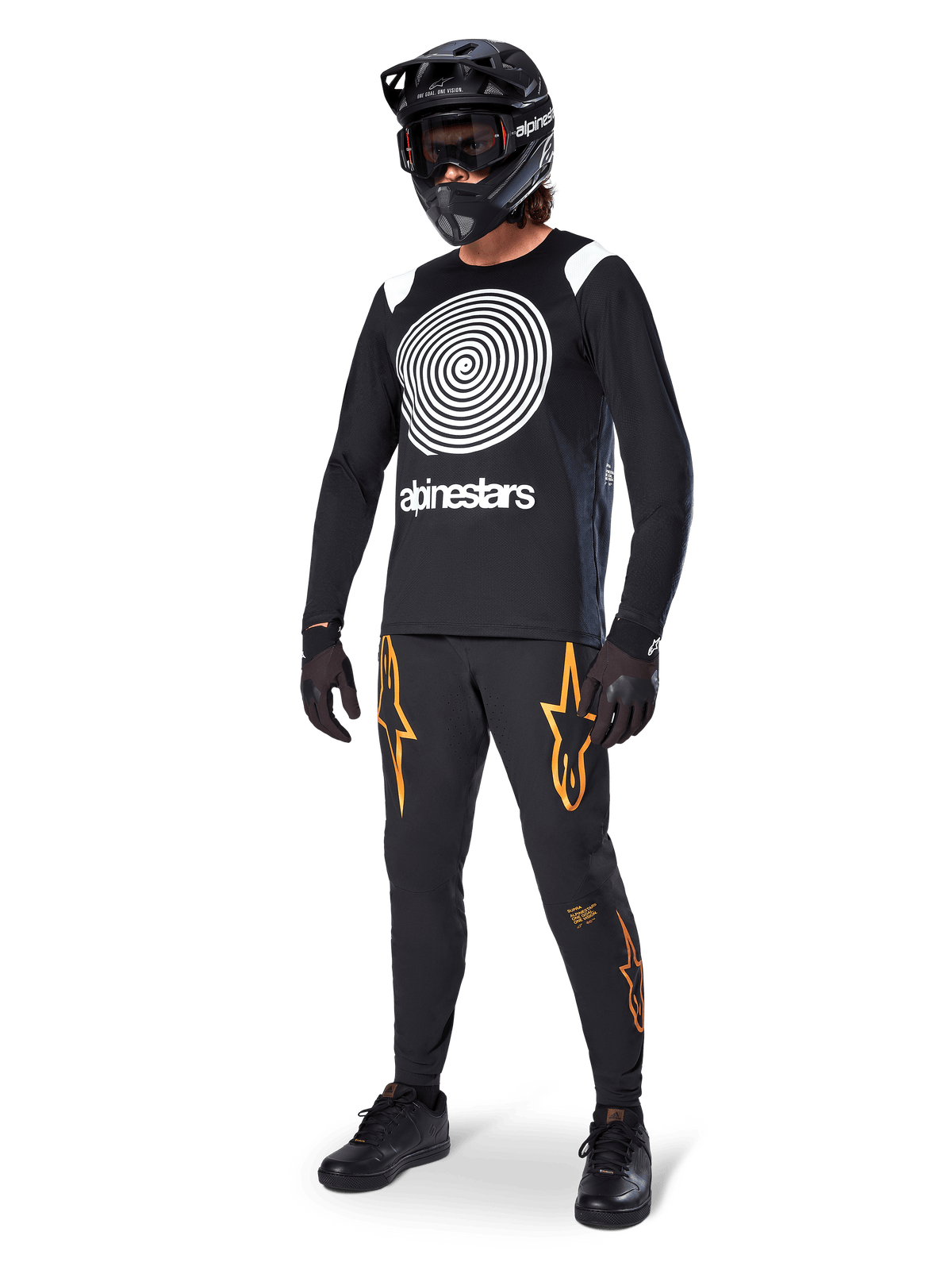 MTB-rijder draagt Alpinestars A-Supra Oscar Jersey met lange mouwen in het zwart, met de witte vintage Oscar-spiraalgrafiek, zwarte broek met oranje sterlogo's, zwarte full-face helm met goggle en zwarte handschoenen, professionele mountainbike-uitrusting