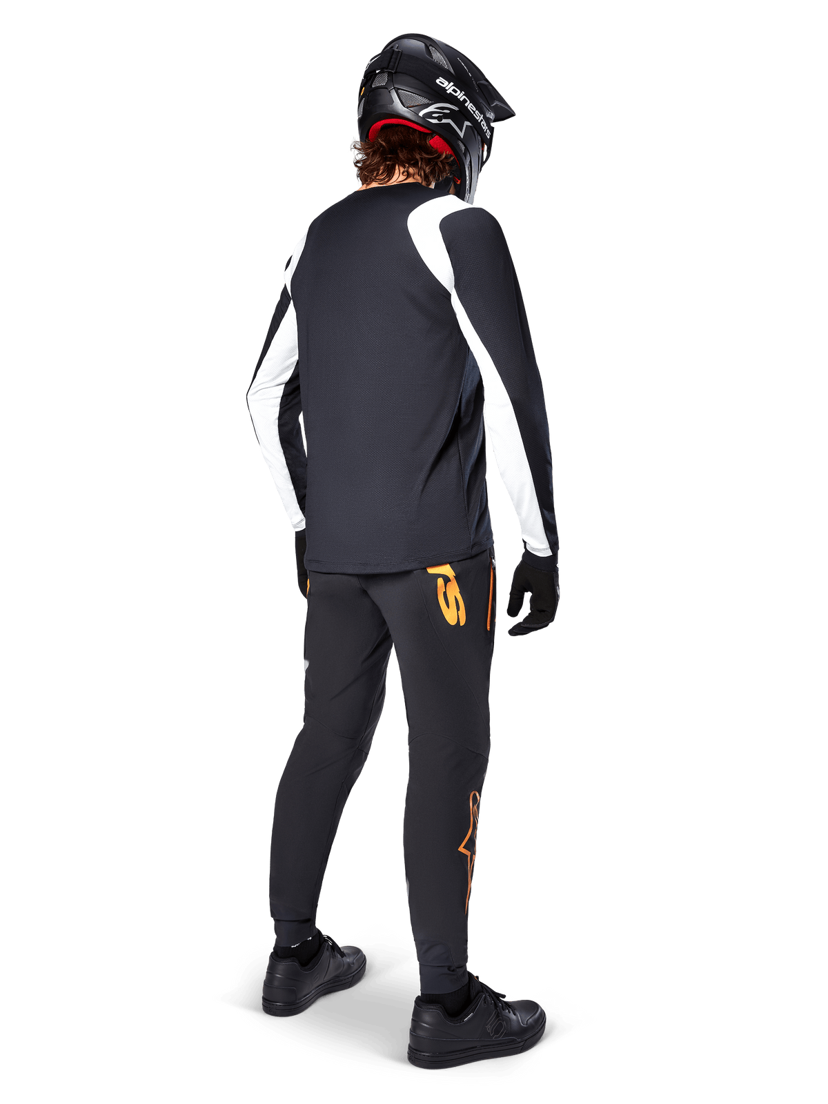 Alpinestars A-Supra Oscar Jersey met lange mouwen, zwart met witte panelen op de mouwen, gedragen door een MTB-rijder van de achterkant gezien met een zwarte full-face helm en zwarte broek met oranje logo's, fietskleding voor off-road gravity riding