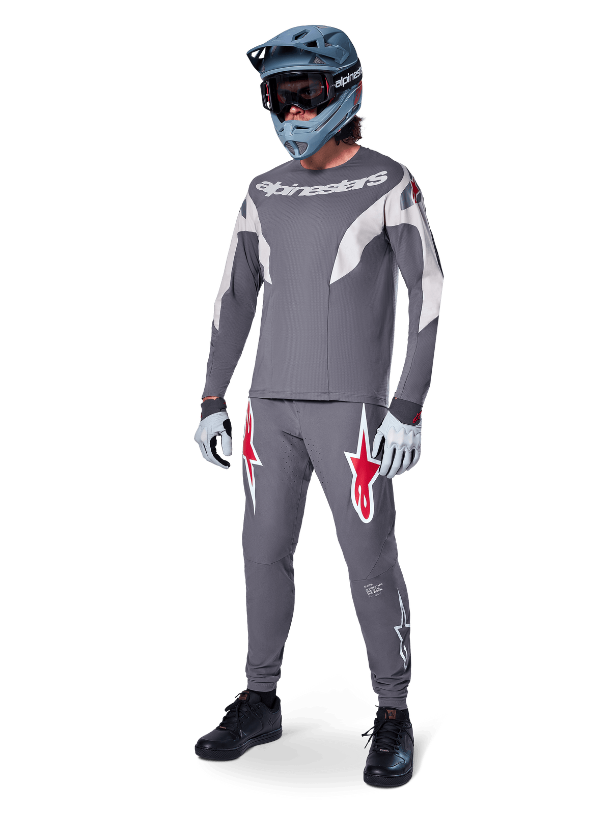 Pilote de VTT portant un maillot Alpinestars A-Supra Race à manches longues, gris foncé avec des accents gris clair, pantalon gris foncé assorti avec logo étoile rouge, casque intégral gris avec lunettes, gants gris clair, tenue coordonnée pour la course de VTT