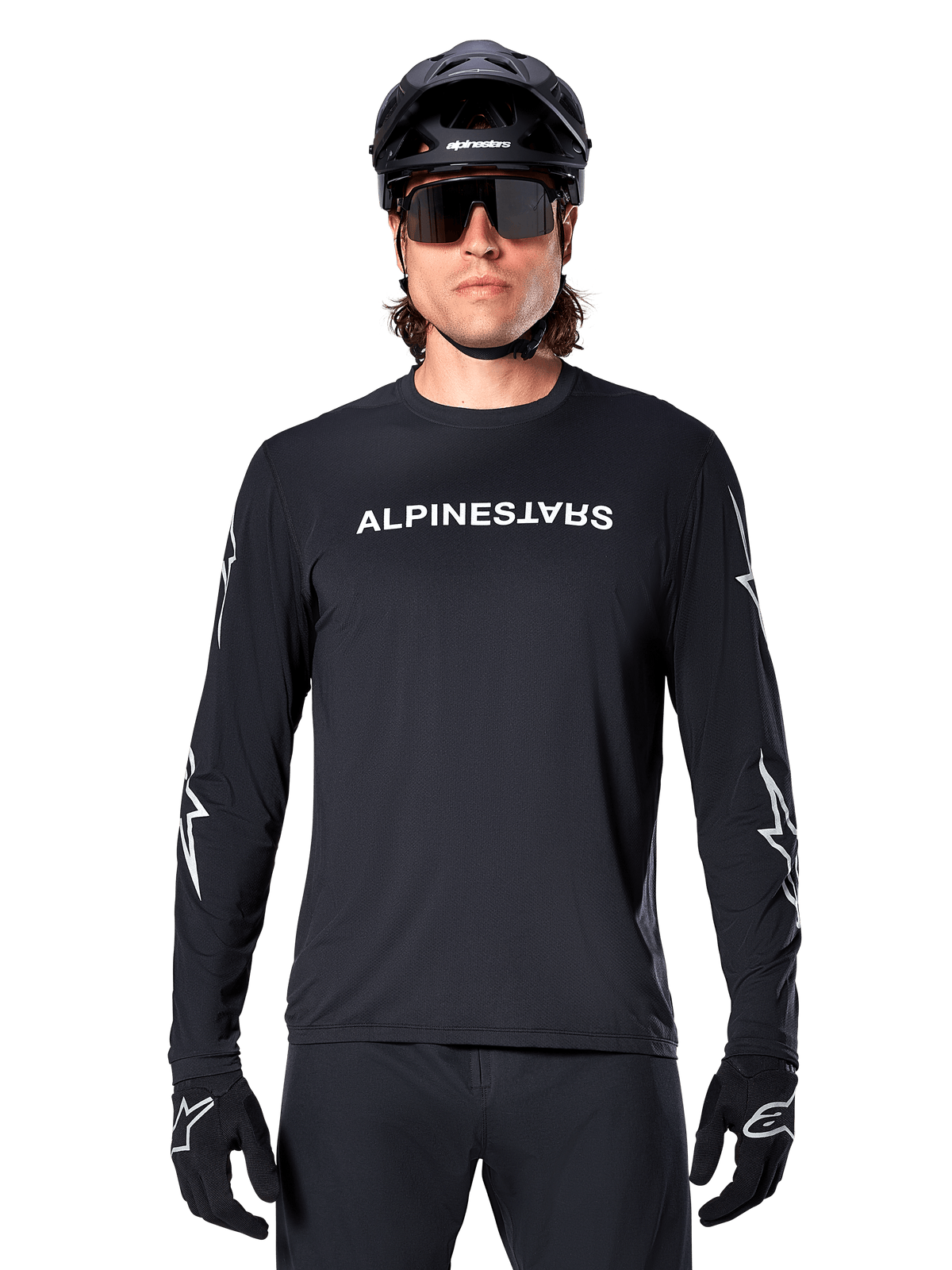 Alpinestars A-Dura Switch Jersey met lange mouwen, zwart MTB-shirt, wit Alpinestars-woordmerk op de borst en ster-graphics op de mouwen, gedragen met een zwarte mountainbikehelm en sportzonnebril, technische kleding voor trailrijden