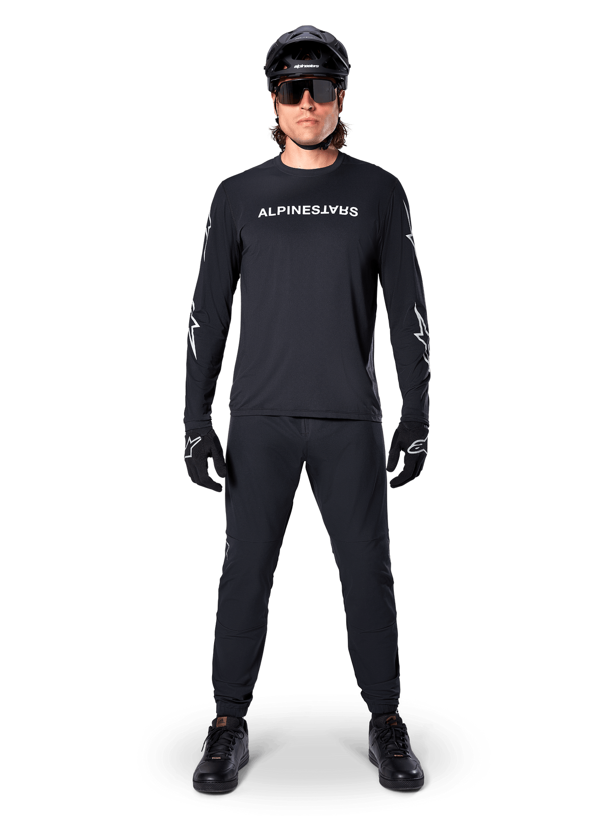 Alpinestars A-Dura Switch Jersey met lange mouwen, zwart, gedragen door een mountainbiker met bijpassende zwarte broek en helm, voorzien van wit woordmerk-logo op de borst, technische off-road fietskleding