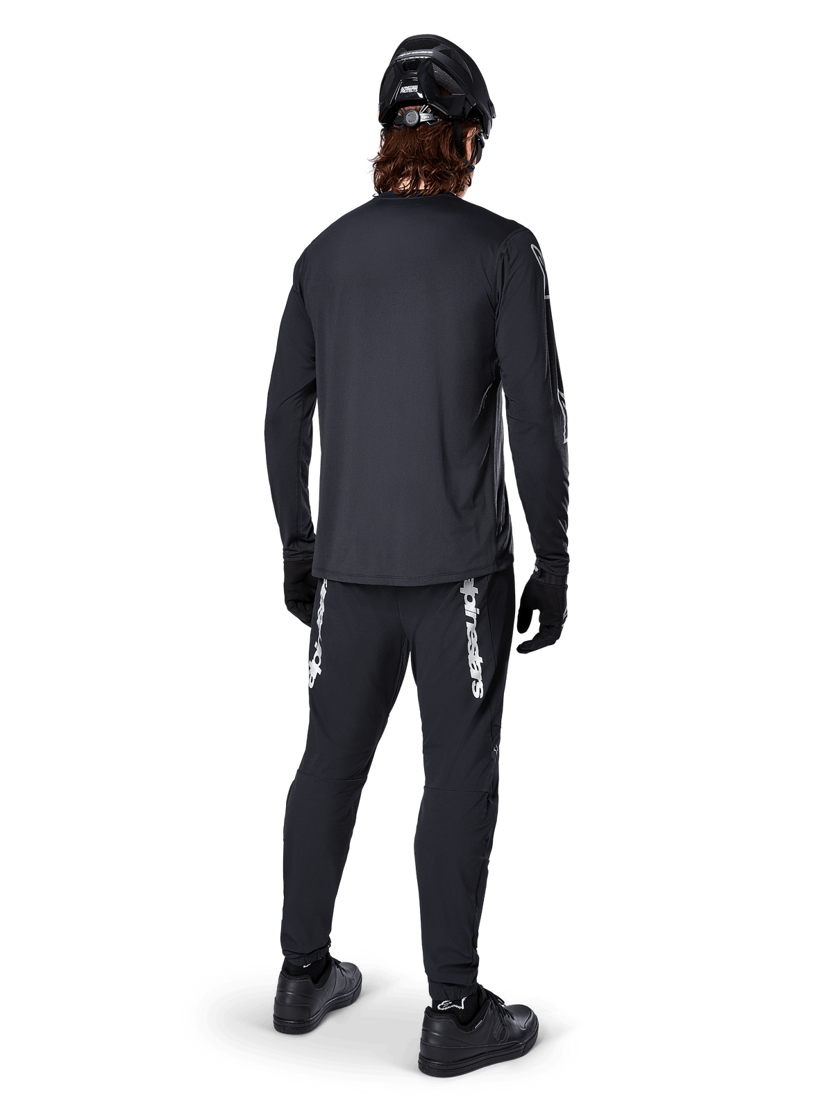 Alpinestars A-Dura Switch Jersey met lange mouwen, zwart, achteraanzicht op een rijder, gedragen met een zwarte broek met witte Alpinestars-branding, zwarte helm en handschoenen, voorzien van een langere drop tail voor extra dekking, technische mountainbikekleding