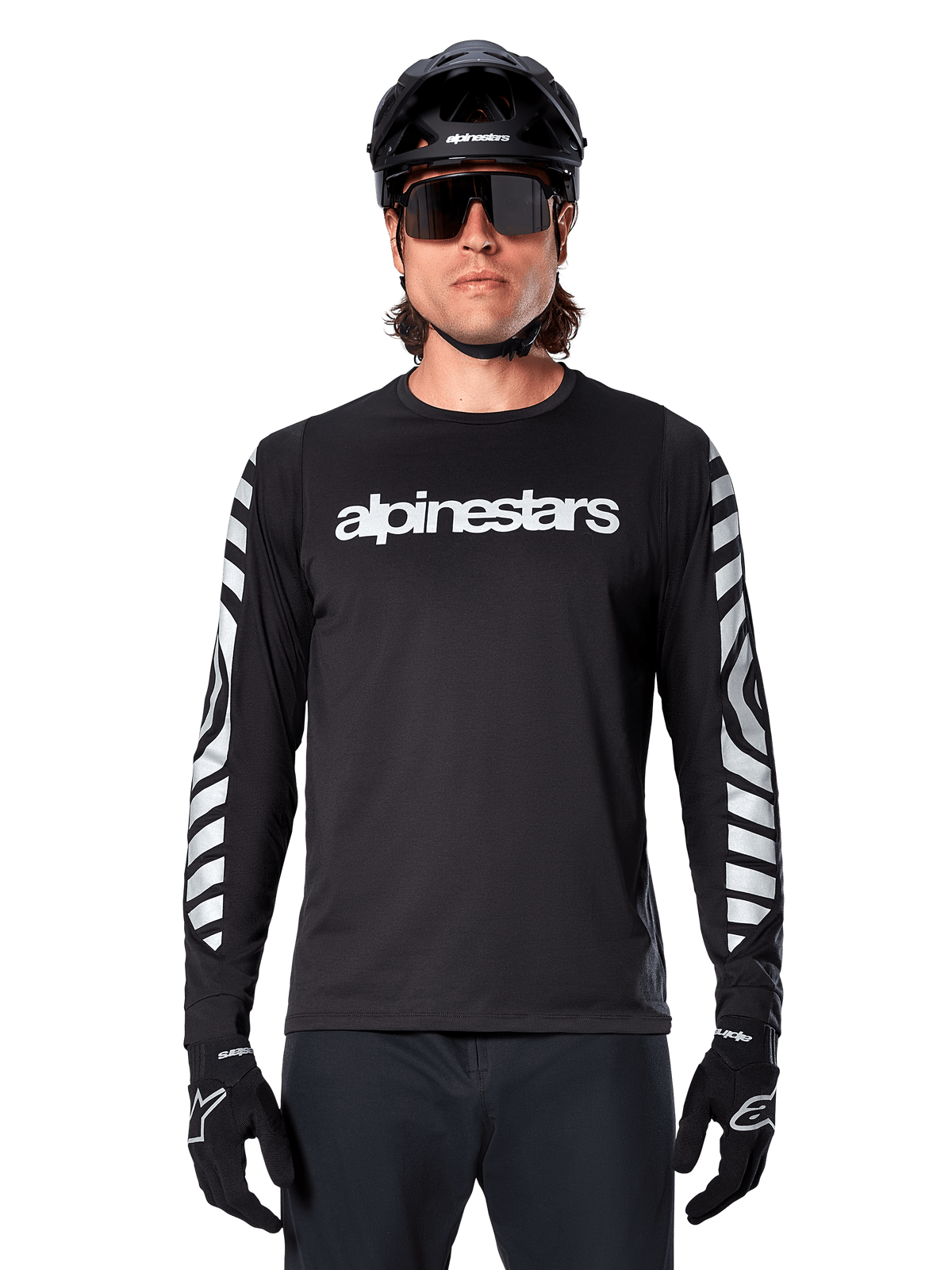 Male rider wearing Alpinestars A-Dura Dri Oscar Long Sleeve Jersey, Black Reflective, featuring white alpinestars wordmark on chest and silver reflective graphics on sleeves, paired with black MTB helmet, sunglasses, and black gloves, performance cycling gear