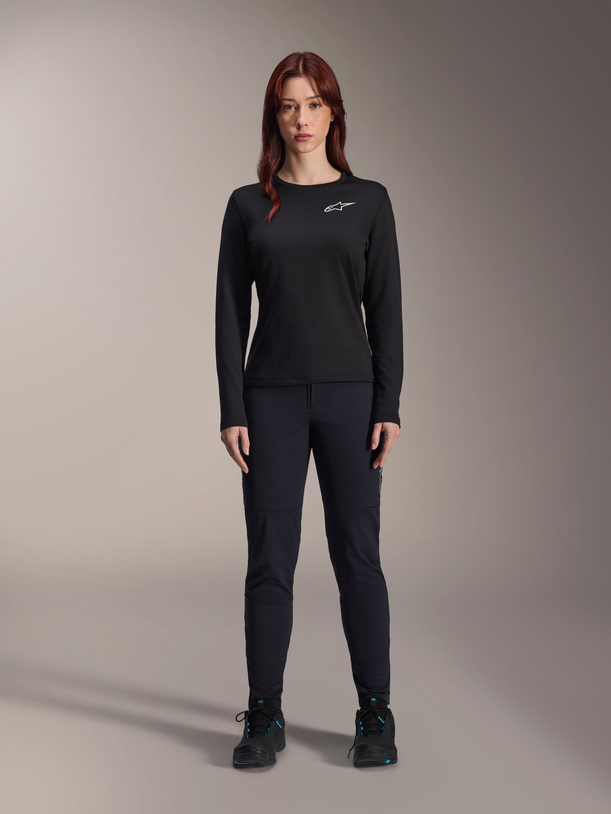 Maillot thermique Stella A-Dura d'Alpinestars, maillot de VTT à manches longues pour femme en noir, avec un logo blanc sur la poitrine, porté avec un équipement noir assorti et des chaussures de cyclisme, portrait en studio.