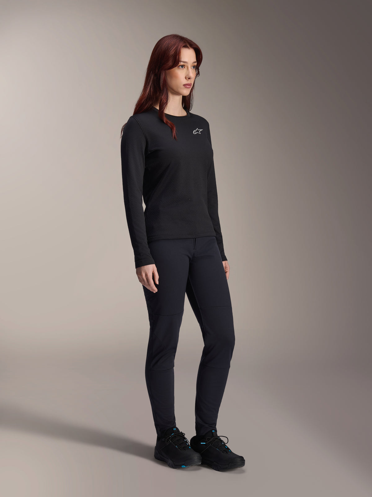 Femme portant le maillot thermique Alpinestars Stella A-Dura, maillot de VTT noir à manches longues avec logo blanc sur la poitrine, associé à un pantalon noir assorti et des chaussures de sport, portrait en studio pour vêtements de cyclisme de performance
