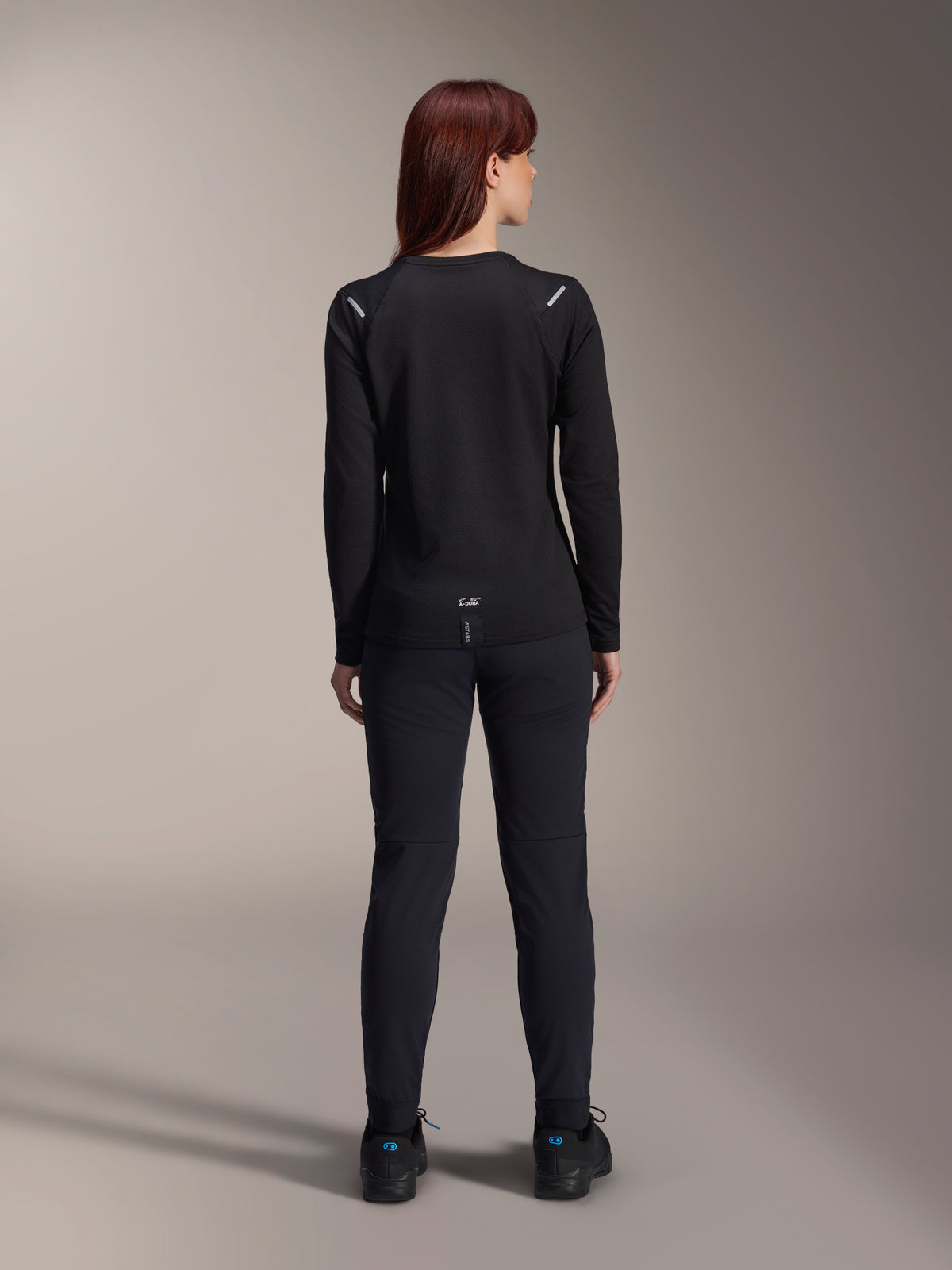 Maillot thermique Stella A-Dura Alpinestars pour femme, vue arrière, noir, maillot de cyclisme à manches longues avec polaire isolante, porté avec un pantalon et des chaussures noirs assortis, doté d'un petit marquage A-Dura en bas du dos et d'accents réfléchissants sur les épaules, conçu pour la pratique du VTT féminin.