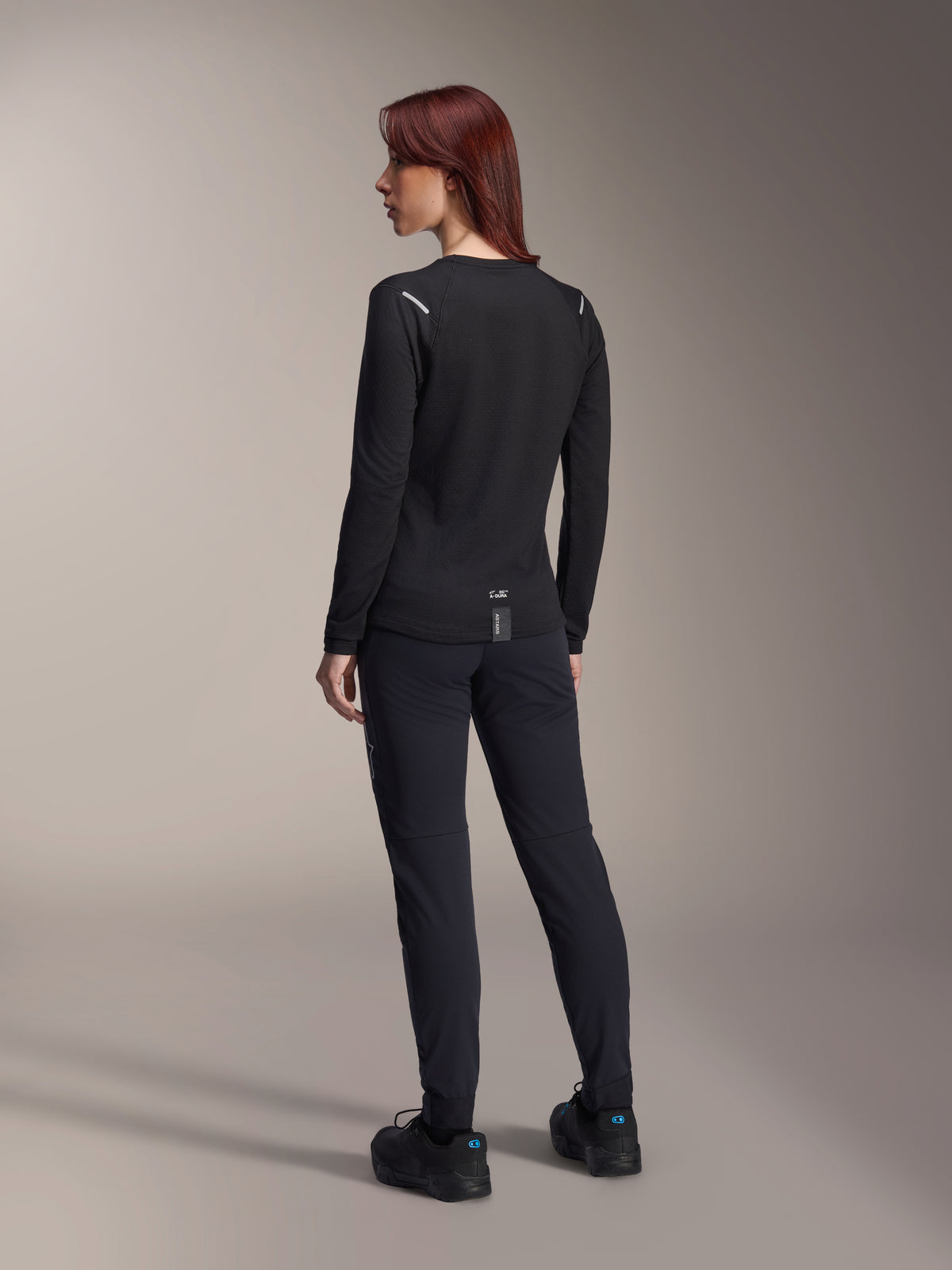 Maillot thermique pour femme Alpinestars Stella A-Dura, maillot de VTT à manches longues noir, vue arrière montrant les détails réfléchissants aux épaules et le pan arrière allongé, porté avec un pantalon et des chaussures noirs, équipement de performance pour le VTT.