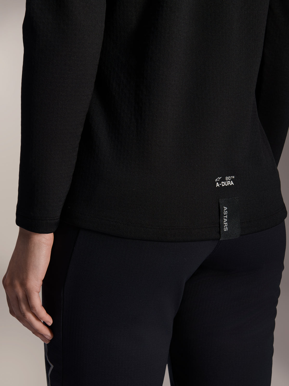 Maillot thermique femme Alpinestars Stella A-Dura, maillot de VTT thermique noir, gros plan sur l'ourlet inférieur arrière avec marquage A-DURA blanc et étiquette logo ASTARS verticale, conçu pour le VTT par temps froid