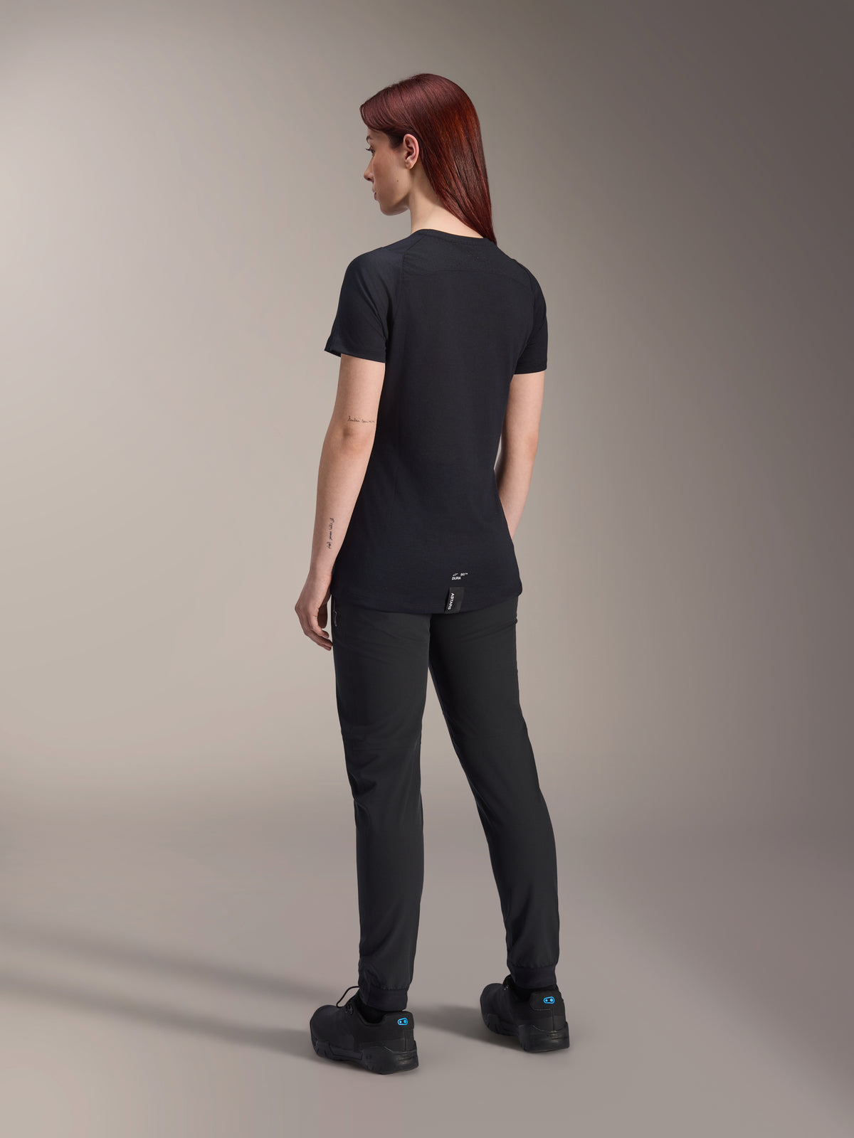 Alpinestars Stella A-Dura Dri Switch Kurzarmtrikot, Schwarz, rothaariges Model in Rückansicht, technische Passform mit verlängertem Rückensaum für optimale Abdeckung beim Radfahren, kombiniert mit schwarzer Mountainbike-Hose und Schuhen