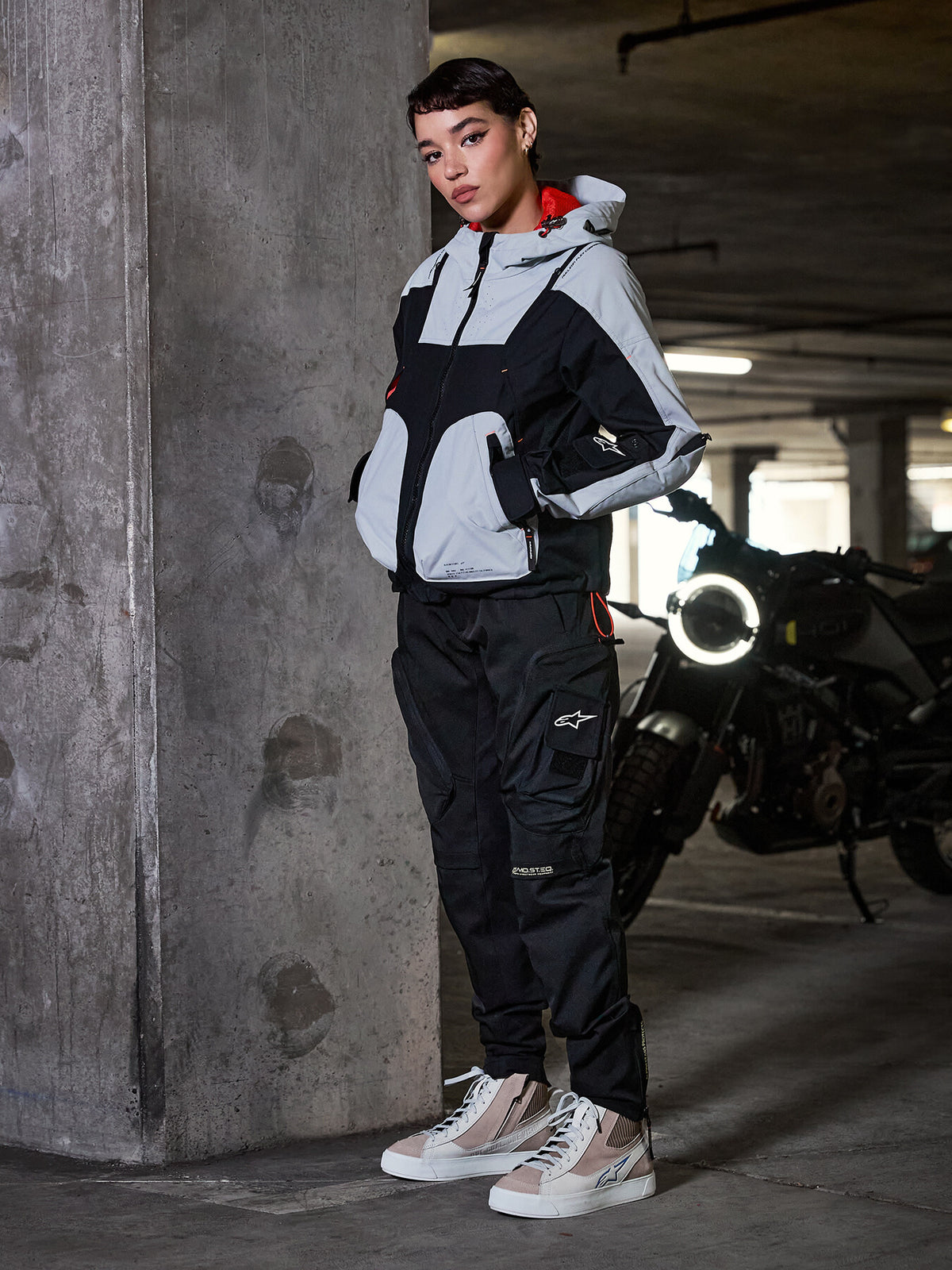 Alpinestars Actea damesbroek, zwart, vrouwelijke motorrijder draagt motorbroek in cargo-stijl met een grijs en zwart technisch jack en beige sneakers, stedelijke parkeergarage-omgeving met motorfiets