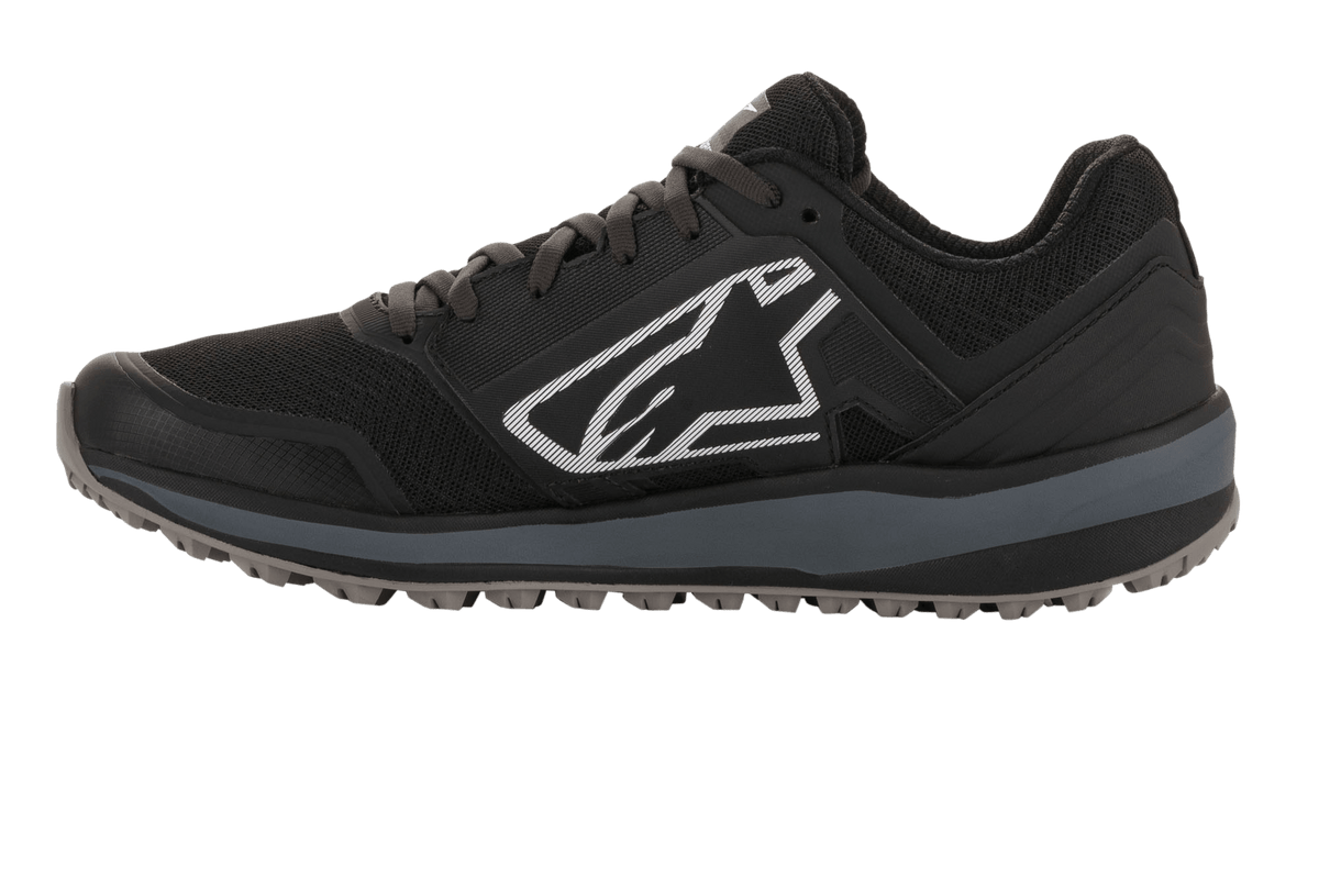 Alpinestars Meta Trail schoenen, performance hardloopschoen, zwart en donkergrijs, zijprofiel met groot wit Astars-logo, donkergrijze tussenzool, oliebestendige rubberen profielzool, ontworpen voor paddockcomfort en gebruik door motorsportteams.