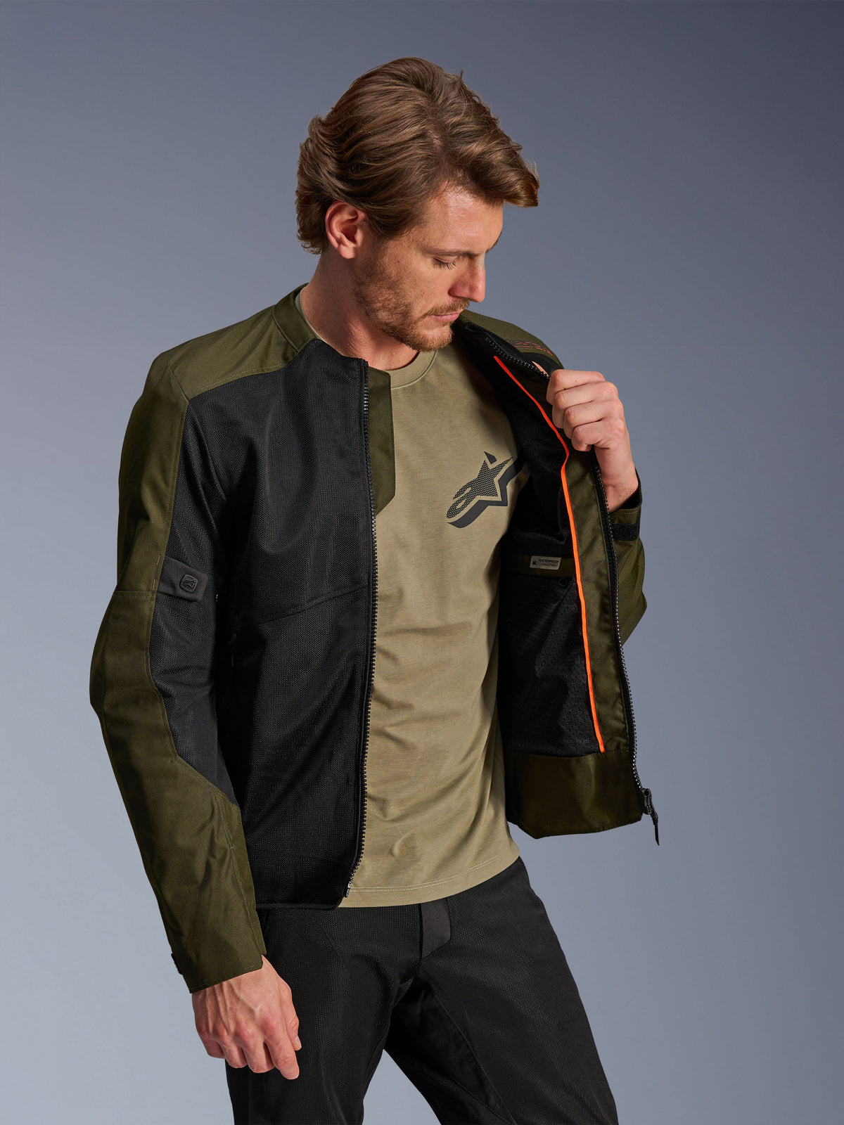 Alpinestars C-1 Air Jacket in Khaki and Black, worn by a male model to show the orange interior lining and waterproof compartment, featuring large tech mesh panels for airflow, urban contemporary motorcycle style paired with a matching olive t-shirt and black pants