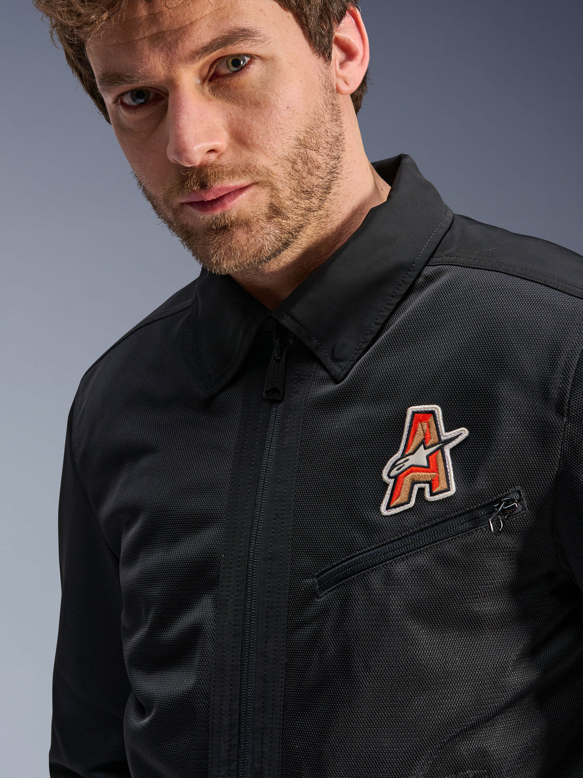 Alpinestars Ignite Air Jacket, zwart, voorzien van een traditionele overhemdkraag en ademende mesh-constructie, met een rood en goud 'A'-logo op de borst, gedragen door een mannelijk model, urbane motorkleding.