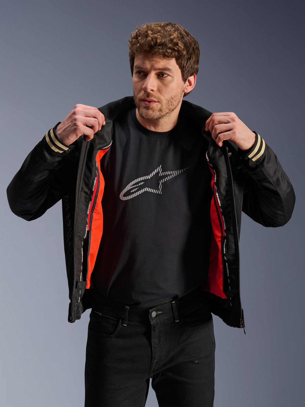 Man die de Alpinestars Ignite Air Jas draagt, zwart, voorzien van een mesh body en feloranje uitneembare winddichte voering, gecombineerd met een zwart t-shirt met Astars-logo en een zwarte broek, urban contemporary motorstijl