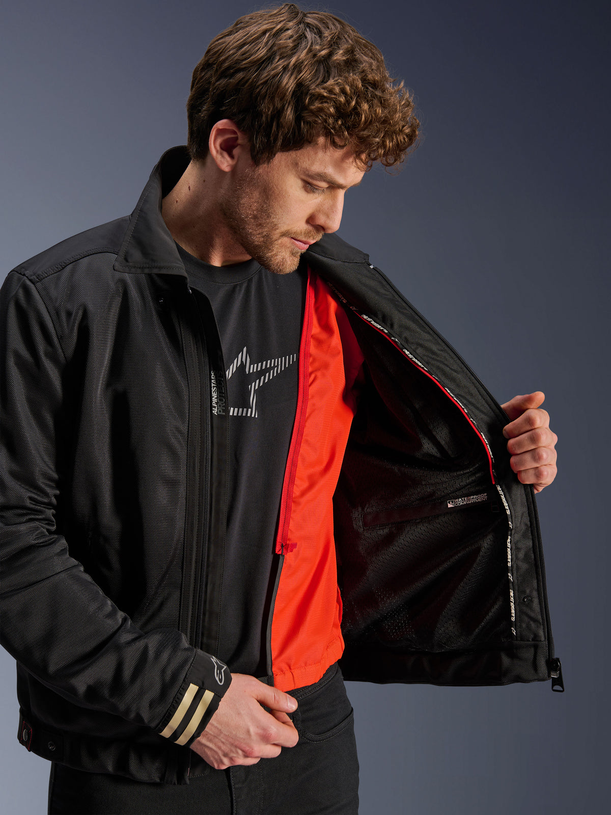 Alpinestars Ignite Air Jacket, zwarte urban motorjas, voorzien van een feloranje afneembare winddichte voering en mesh body, binnenaanzicht met waterdicht compartiment, gedragen met een zwart Alpinestars logo T-shirt.