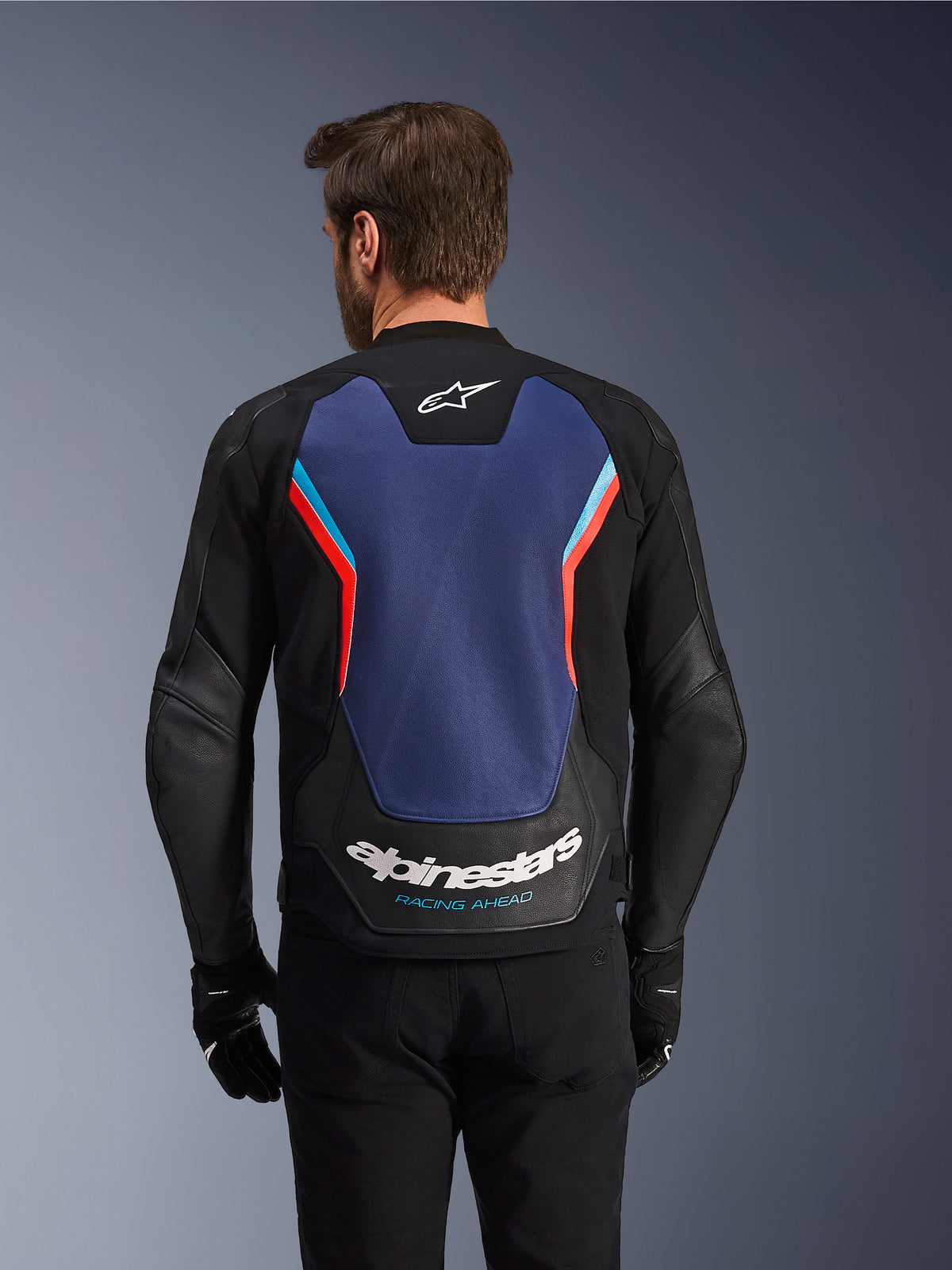 Rückansicht der Alpinestars GP Force V2 Lederjacke in Schwarz, Blau und Red Fluo, mit einem großen blauen Rückenpanel mit fluoreszierenden roten und hellblauen Akzenten, weißem Alpinestars-Logo und „Racing Ahead“-Schriftzug am unteren Rücken, getragen von einem Fahrer mit schwarzen Handschuhen und Hosen.
