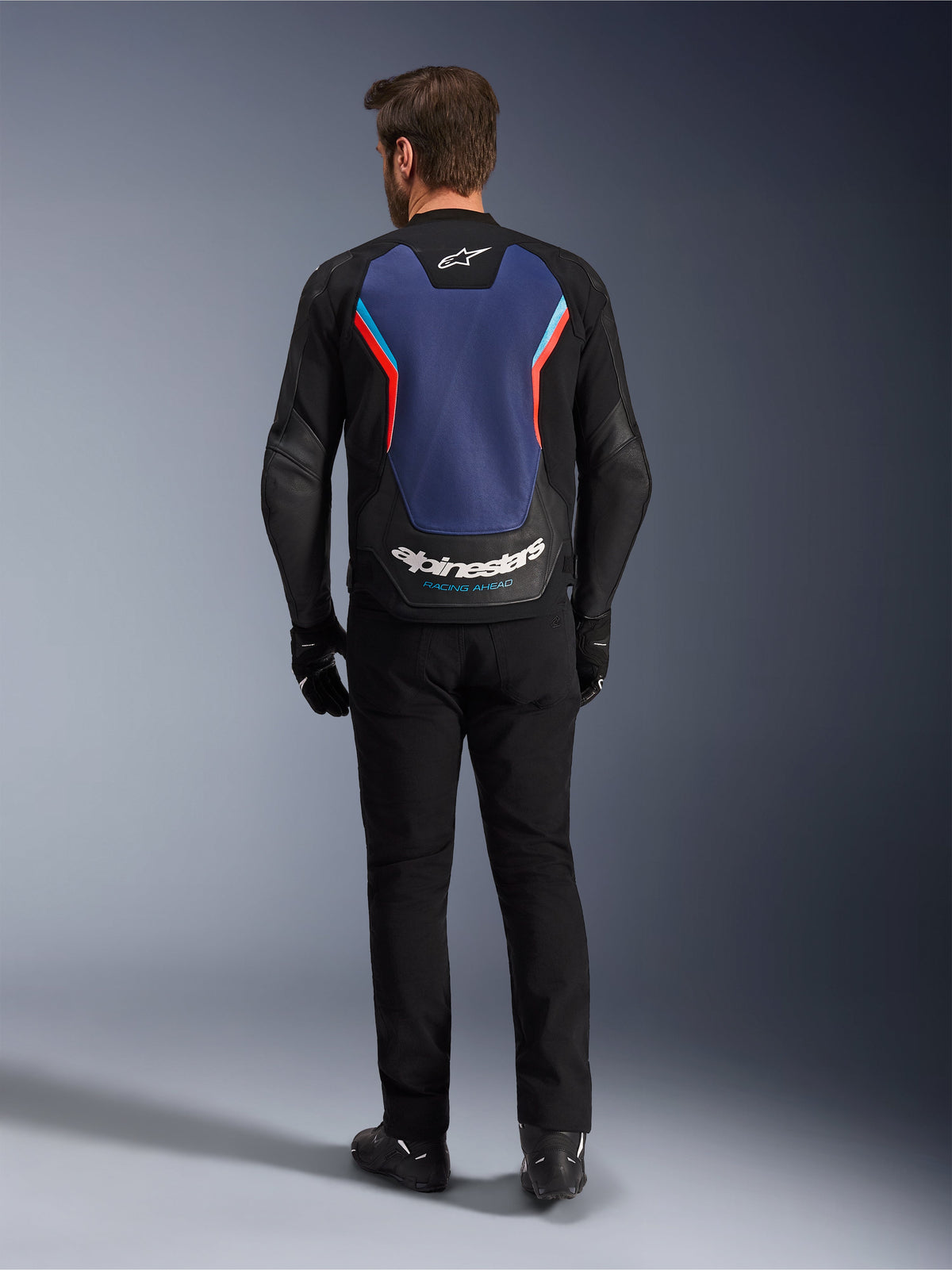 Alpinestars GP Force V2 lederen jas, sport motorjas, zwart blauw rood fluo, achteraanzicht op een model, voorzien van een groot blauw rugpaneel met rode en lichtblauwe accenten en witte Alpinestars-branding op de onderrug, gecombineerd met een zwarte motorbroek en handschoenen.