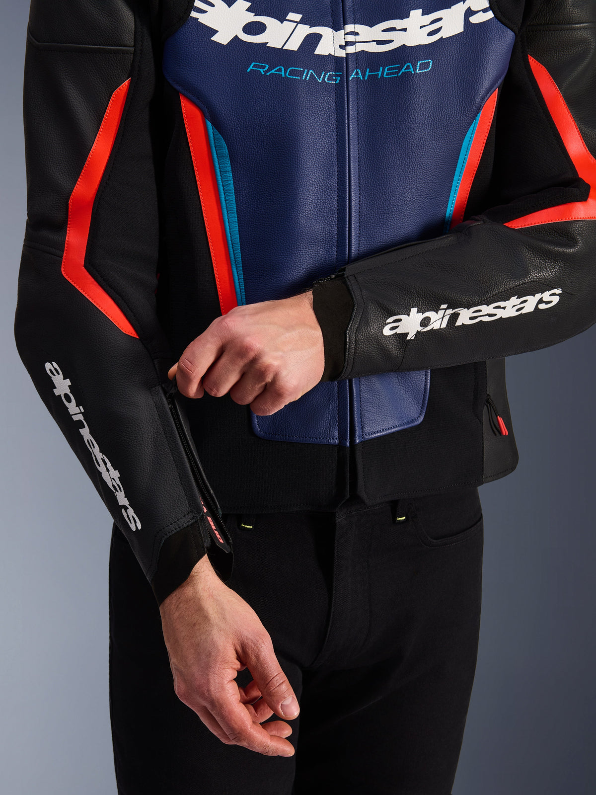 Alpinestars GP Force V2 Leren Jas in zwart, blauw en fluo rood, close-up van een persoon die de rits bij de mouwmanchet verstelt, met witte Alpinestars-logo's op de onderarm en borst, getextureerd rundleer voor sportief motorrijden