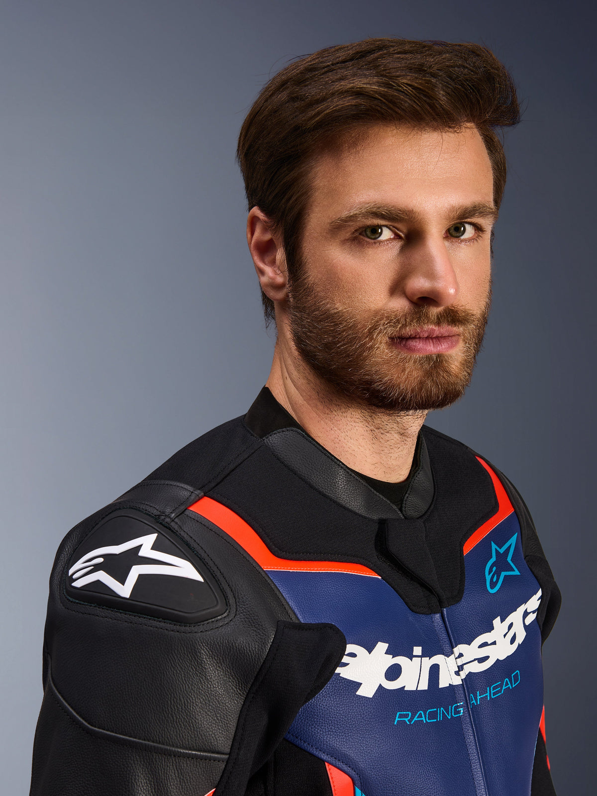 Alpinestars GP Force V2 lederen motorjas, zwart blauw en fluorood, close-up portret van een mannelijke motorrijder, met wit Alpinestars-logo op zwarte DFS-schouderbeschermers, blauw lederen borstpaneel met witte branding en 'Racing Ahead'-tekst, fluorode accenten, hoogwaardige sportieve motoruitrusting
