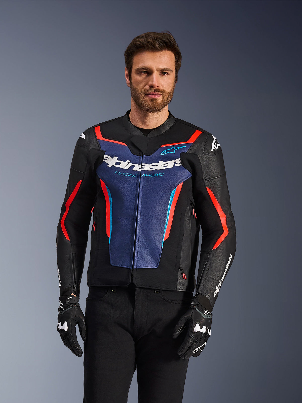 Alpinestars GP Force V2 leren jas, zwart, blauw en fluo rood, gedragen door een mannelijk model met zwarte handschoenen en broek, blauw borstpaneel met 'Alpinestars Racing Ahead' tekst, rode accenten en externe schouderbeschermers, sportieve beschermende motorkleding