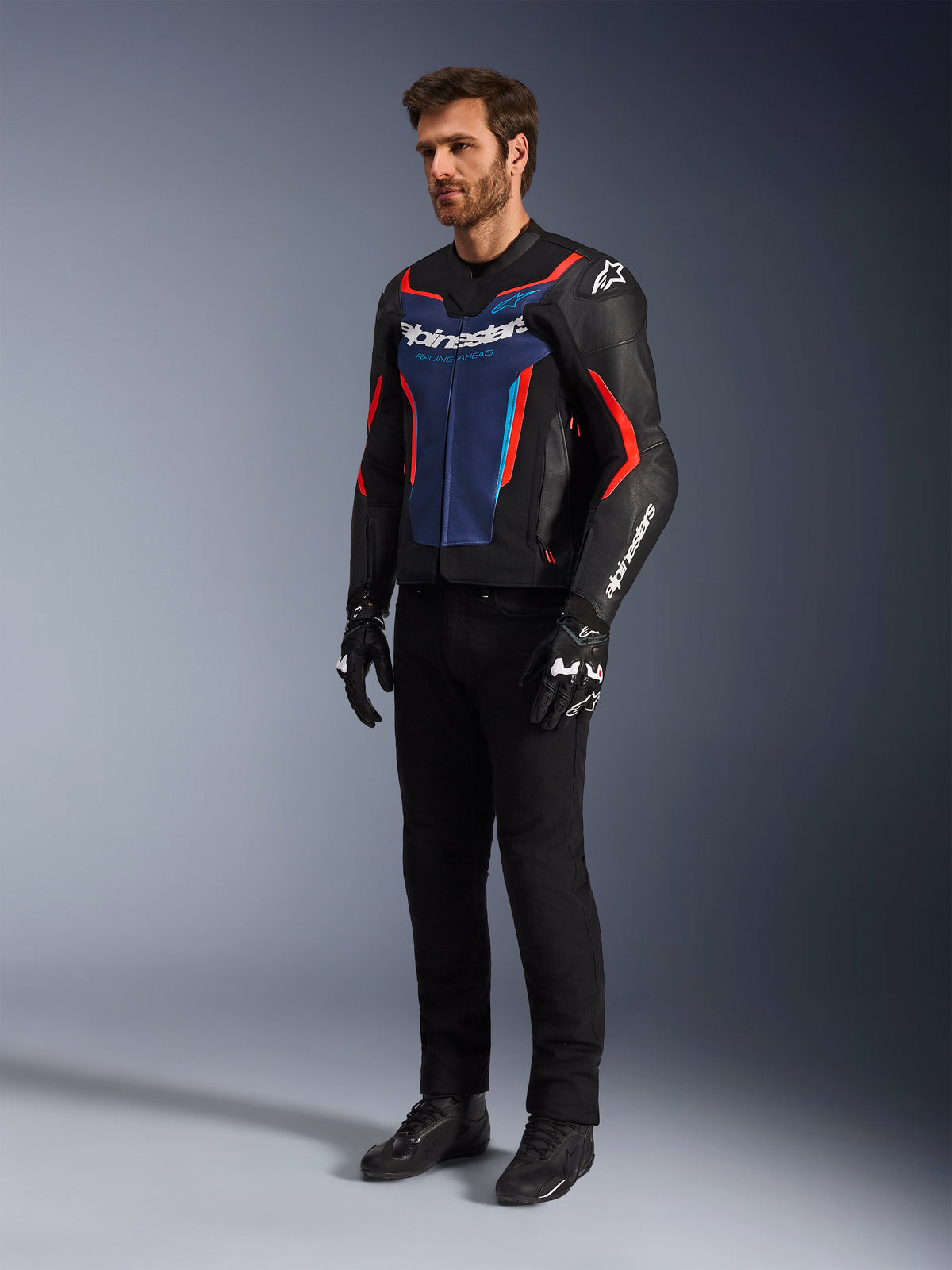 Model draagt de Alpinestars GP Force V2 leren jas in Black Blue Red Fluo, gecombineerd met een zwarte technische motorbroek, zwarte handschoenen en laarzen, professionele motorrace-uitrusting voor sportief rijden