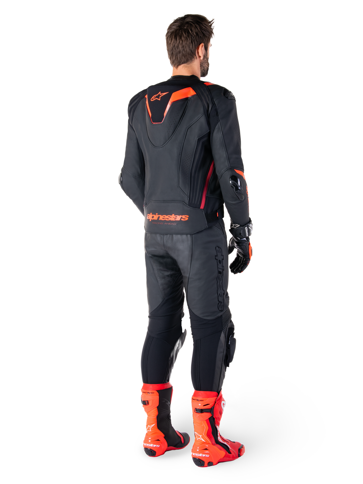 Alpinestars Missile V3 Ignition leren jas, race-sportmotorjas, zwart en fluo rood, achteraanzicht van een rijder die een volledige race-outfit draagt met bijpassende leren broek, handschoenen en rode laarzen, voorzien van een sport Aero-hump en opvallende merknaam op de onderrug.