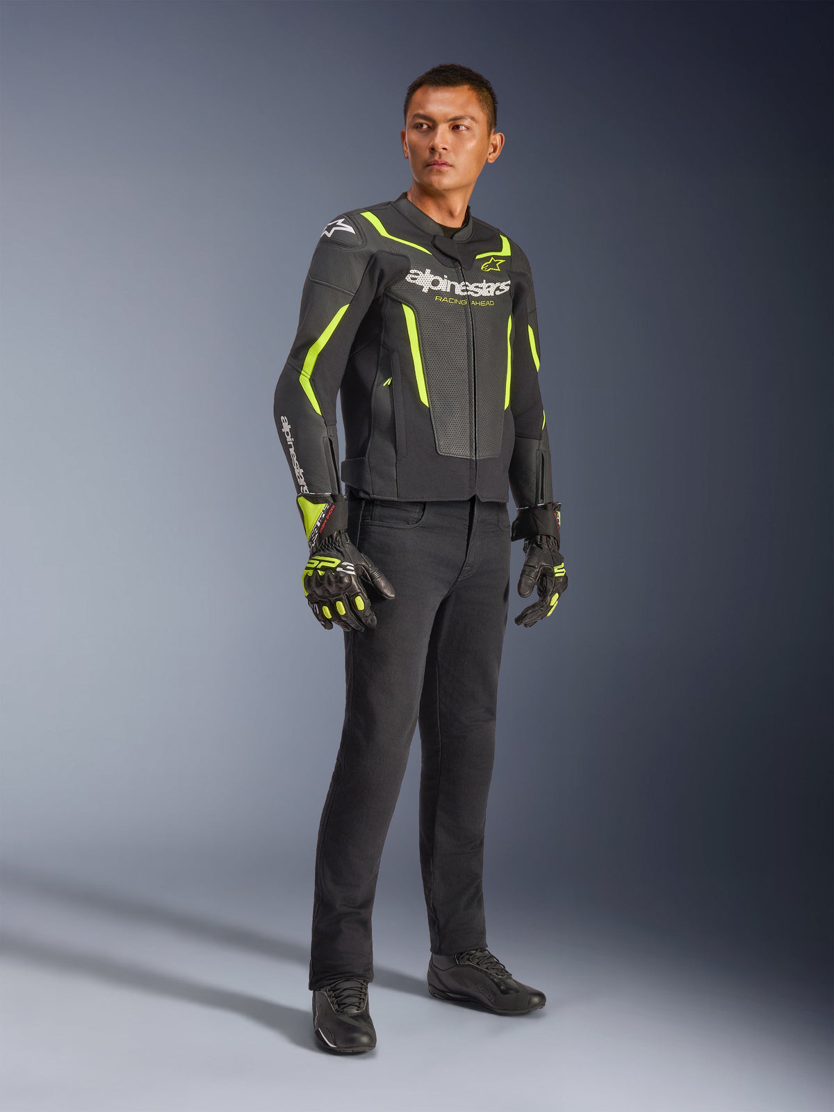 Männliches Model in Alpinestars GP Force V2 Air Lederjacke in Schwarz und Gelb Fluo, kombiniert mit schwarzer Motorradhose, passenden Handschuhen und schwarzen Schuhen, Ganzkörper-Studioaufnahme von sportlicher Motorradausrüstung mit perforiertem Leder und markantem Branding