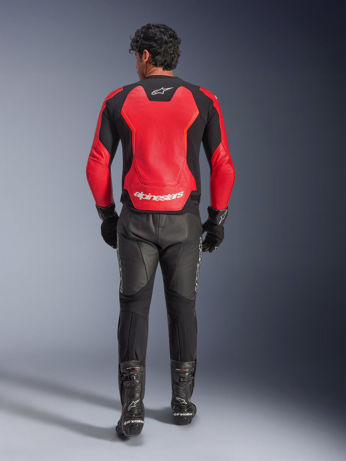 Vue arrière d'un pilote portant le blouson en cuir Alpinestars GP Force V2 Air en rouge et noir, doté de larges panneaux perforés et du logo Alpinestars blanc au dos, accompagné d'un pantalon de moto en cuir noir et de bottes de course.