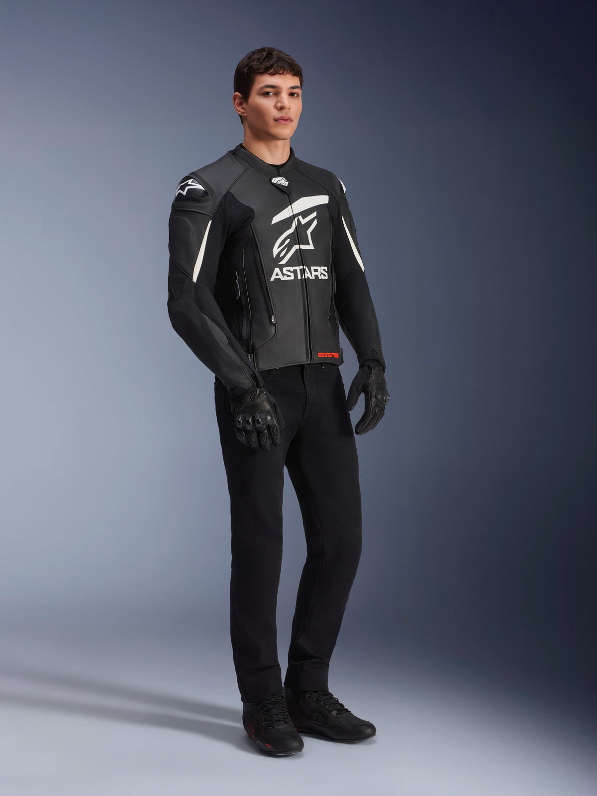 Alpinestars GP Plus V4 Lederjacke, Sport-Motorradjacke, Schwarz und Weiß, getragen von einem männlichen Model mit schwarzer Motorradhose und Handschuhen, großes weißes ASTARS-Logo auf der Brust, externe DFS-Schulterschleifer, professionelles vom Rennsport abgeleitetes Design