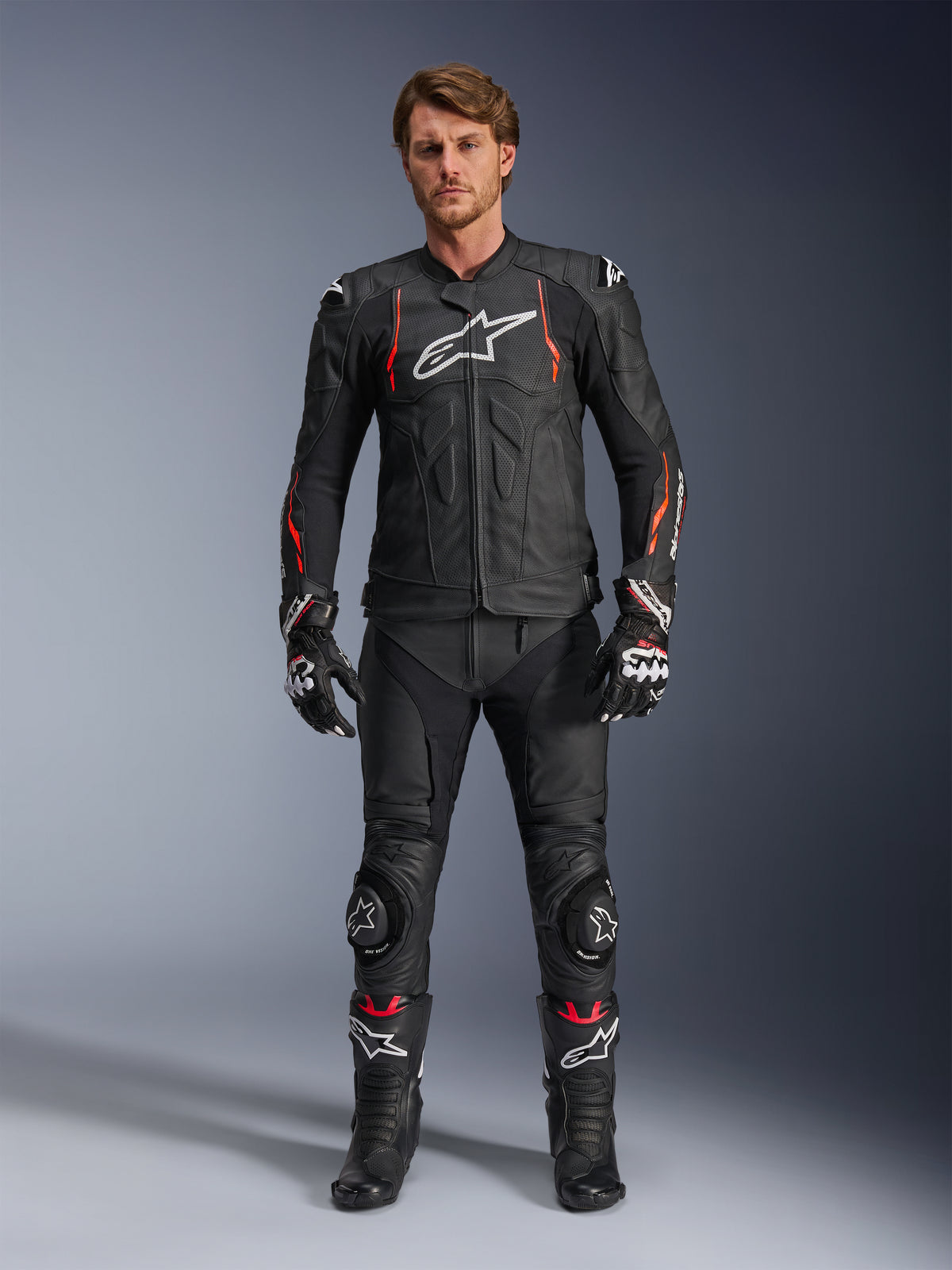 Mannelijke motorrijder in de Alpinestars Dusk Airflow leren jas in Black Red Fluo, voorzien van geperforeerde leren panelen en een wit logo op de borst, gecombineerd met bijpassende zwarte leren motorbroek, kneesliders, handschoenen en racelaarzen, complete motoruitrusting.