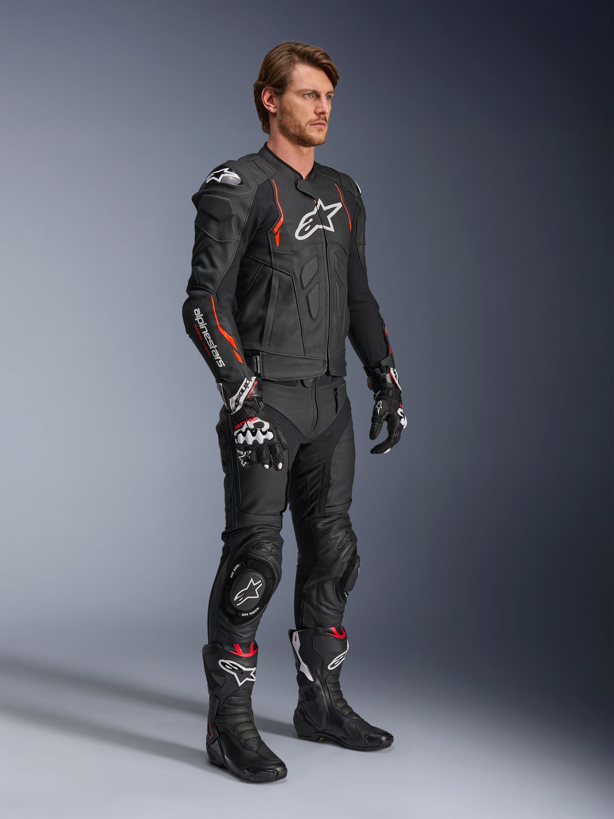 Alpinestars Dusk Airflow Leren Jas, Black Red Fluo, gedragen als onderdeel van een complete motorrace-outfit met bijpassende leren broek, handschoenen en laarzen, voorzien van een geperforeerde leren constructie en sportieve accenten.